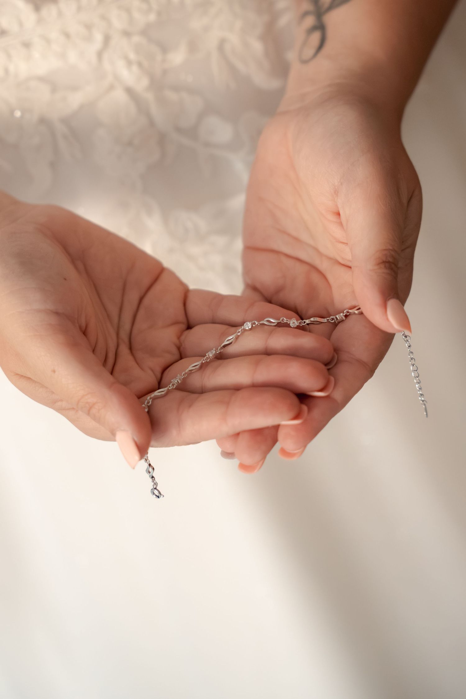 Bride in a lace wedding dress holds a delicate silver twisted chain bracelet in her cupped hands, with a tattoo visible on her wrist.