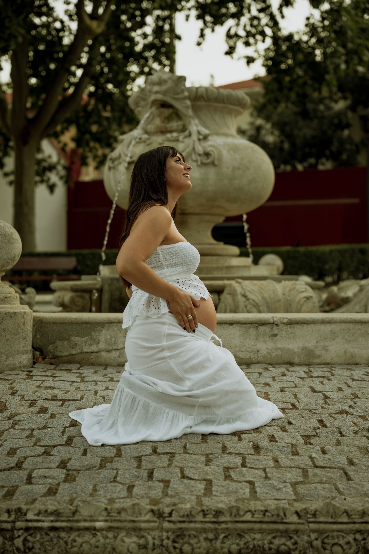 Serene pregnant woman in elegant white dress sitting by stone fountain in outdoor garden setting.