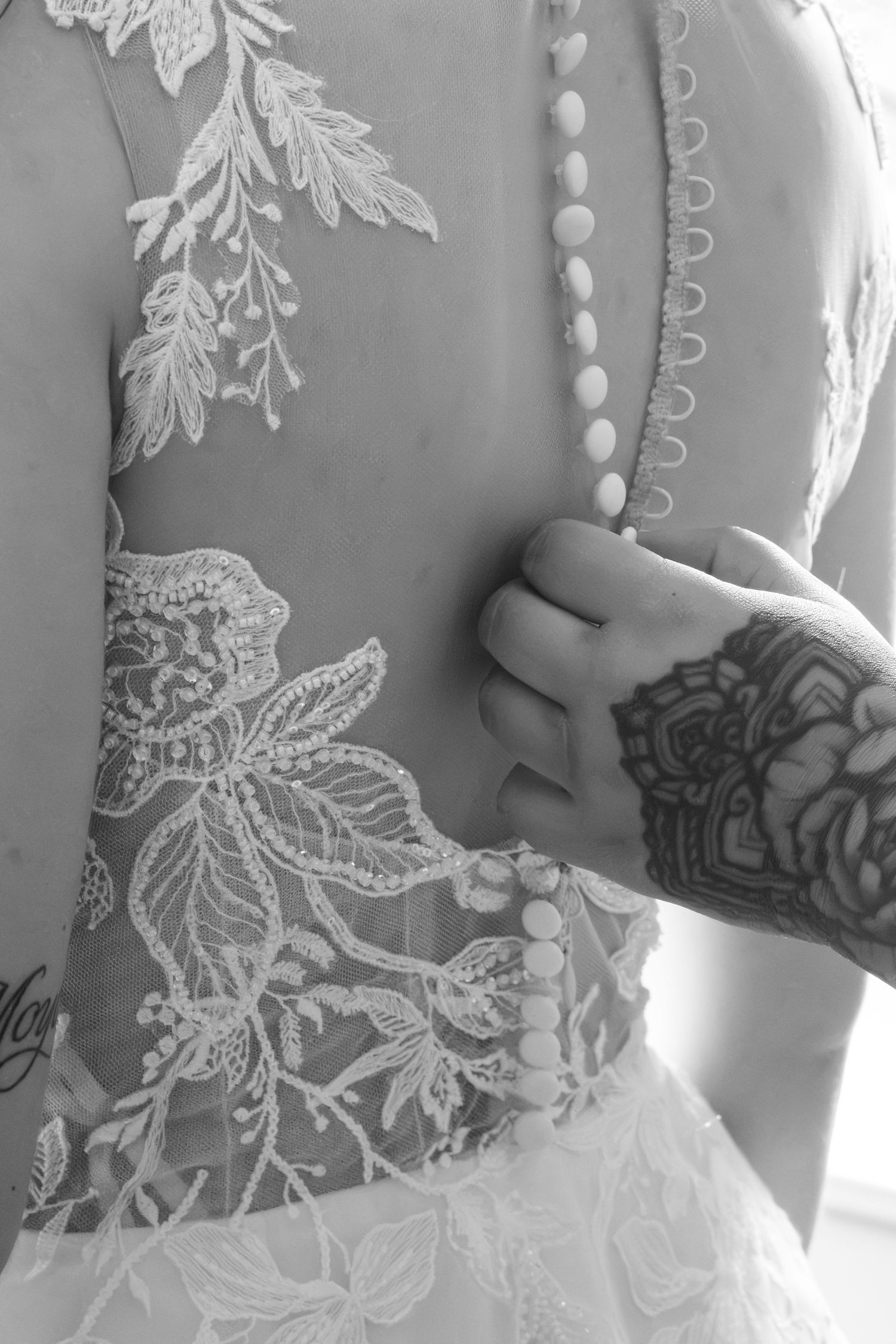 Black and white photo of a tattooed hand buttoning the back of a lace wedding dress with floral appliqué and small covered buttons.