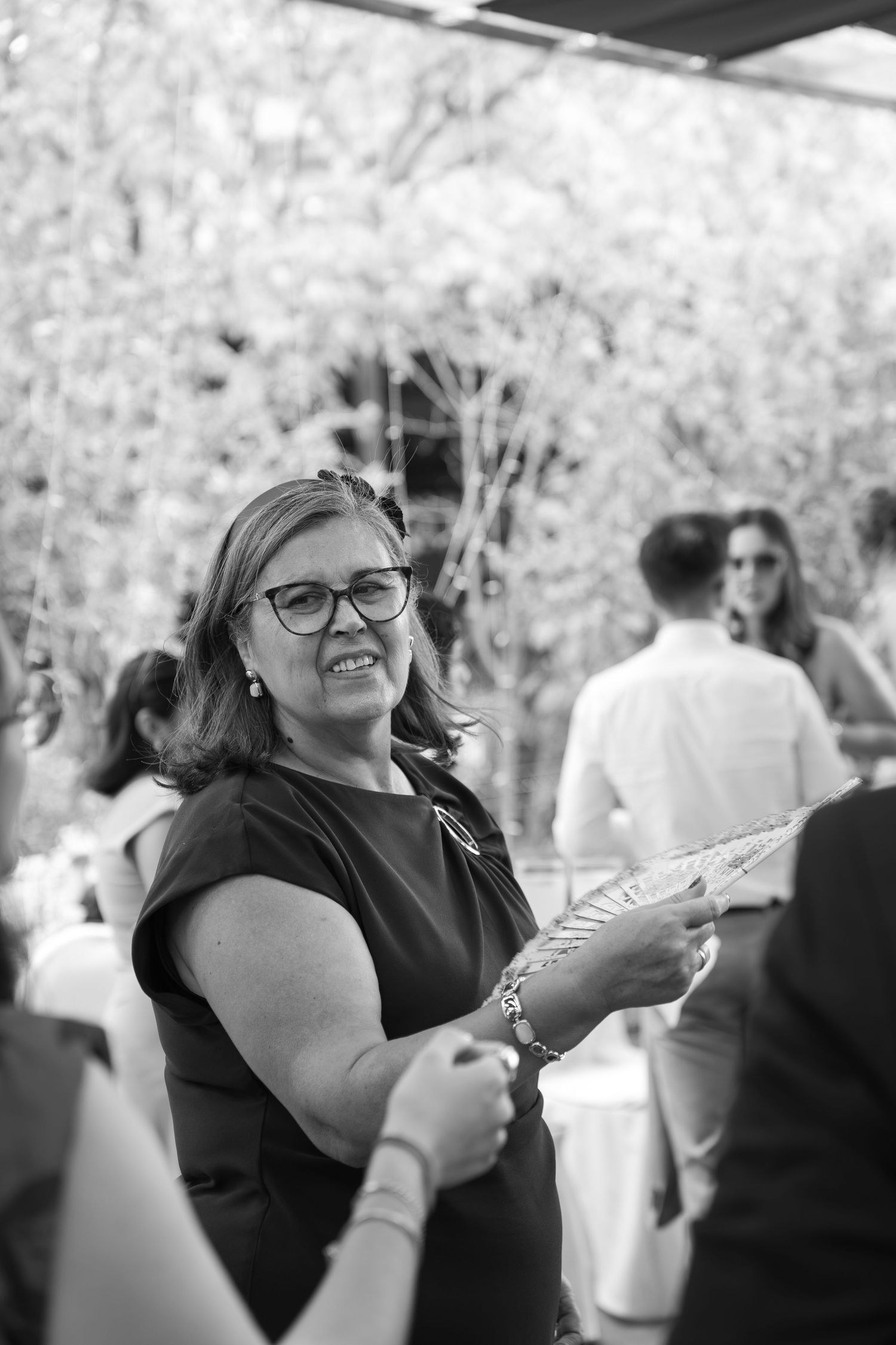 Smiling woman with glasses and a dark top holds a decorative fan at an outdoor gathering in black and white, with guests behind her.