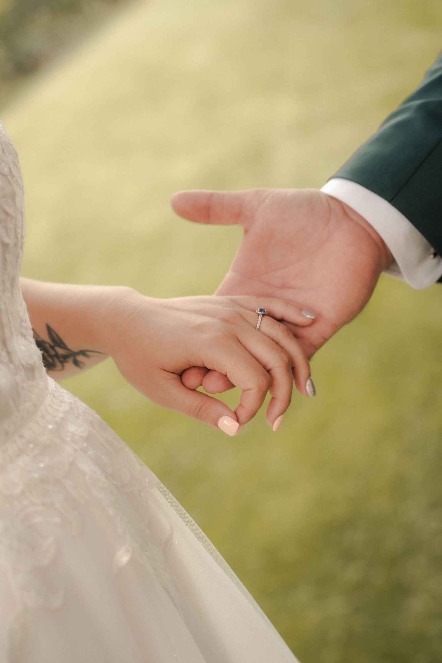 Bride in a white lace gown holds hands with a groom in a green suit, showing her engagement ring against a soft green grassy background.