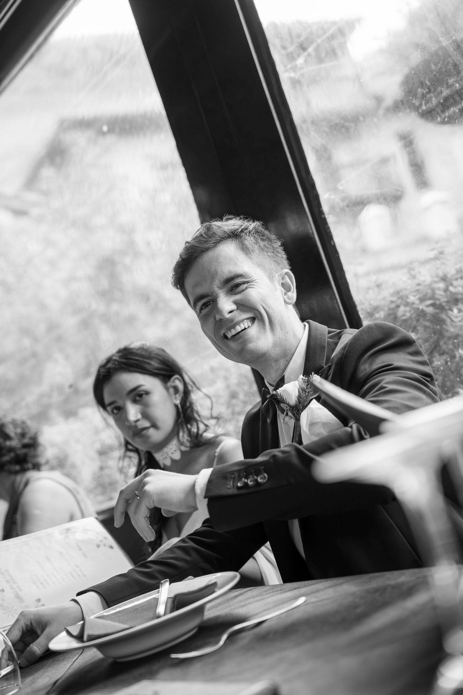 Black and white photo of a smiling man in a suit and bow tie at a restaurant table with a young woman seated beside him by a window.