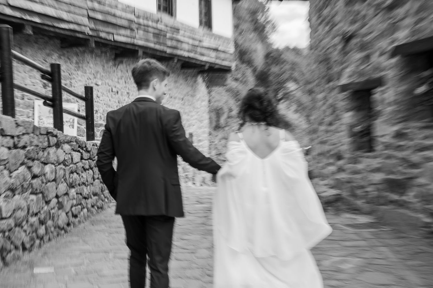 Black and white photo of a groom in a dark suit holding hands with a bride in a white dress as they walk down a stone alley.