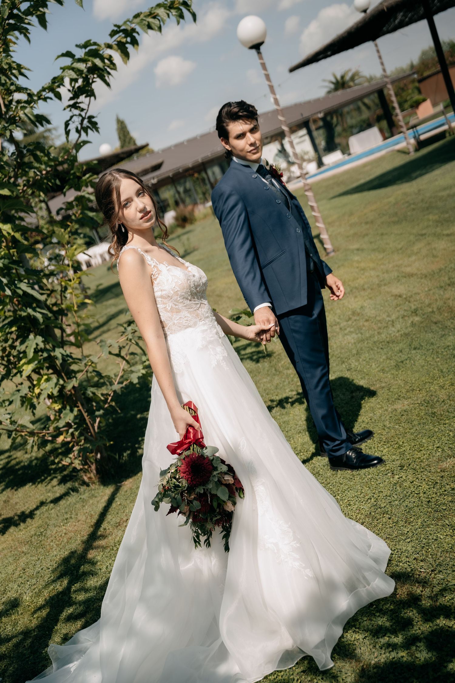 Bride in a lace white gown holding a red bouquet walks hand in hand with a groom in a navy suit across a sunlit garden lawn.