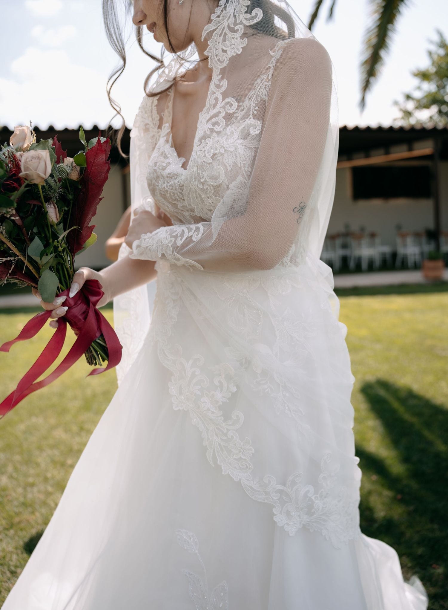 Bride in a lace wedding gown with sheer sleeves and veil holds a bouquet of roses with red ribbons on a sunny garden lawn.
