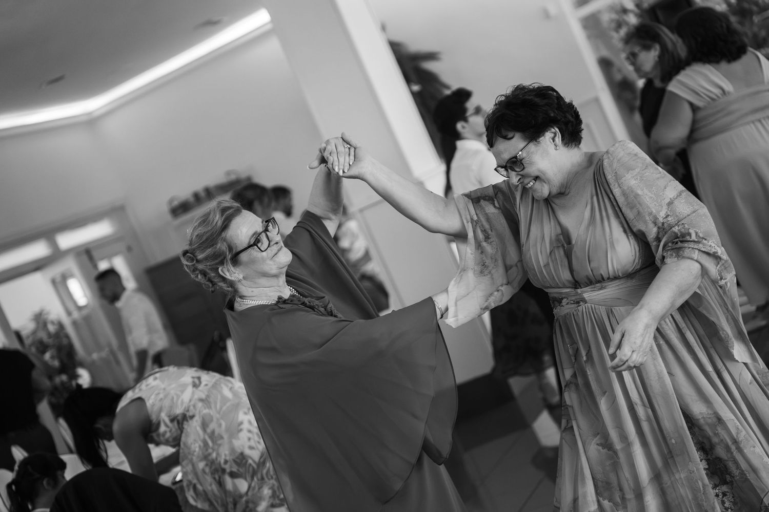 Two smiling women in flowing dresses dance joyfully together holding hands at a lively indoor celebration while guests look on.