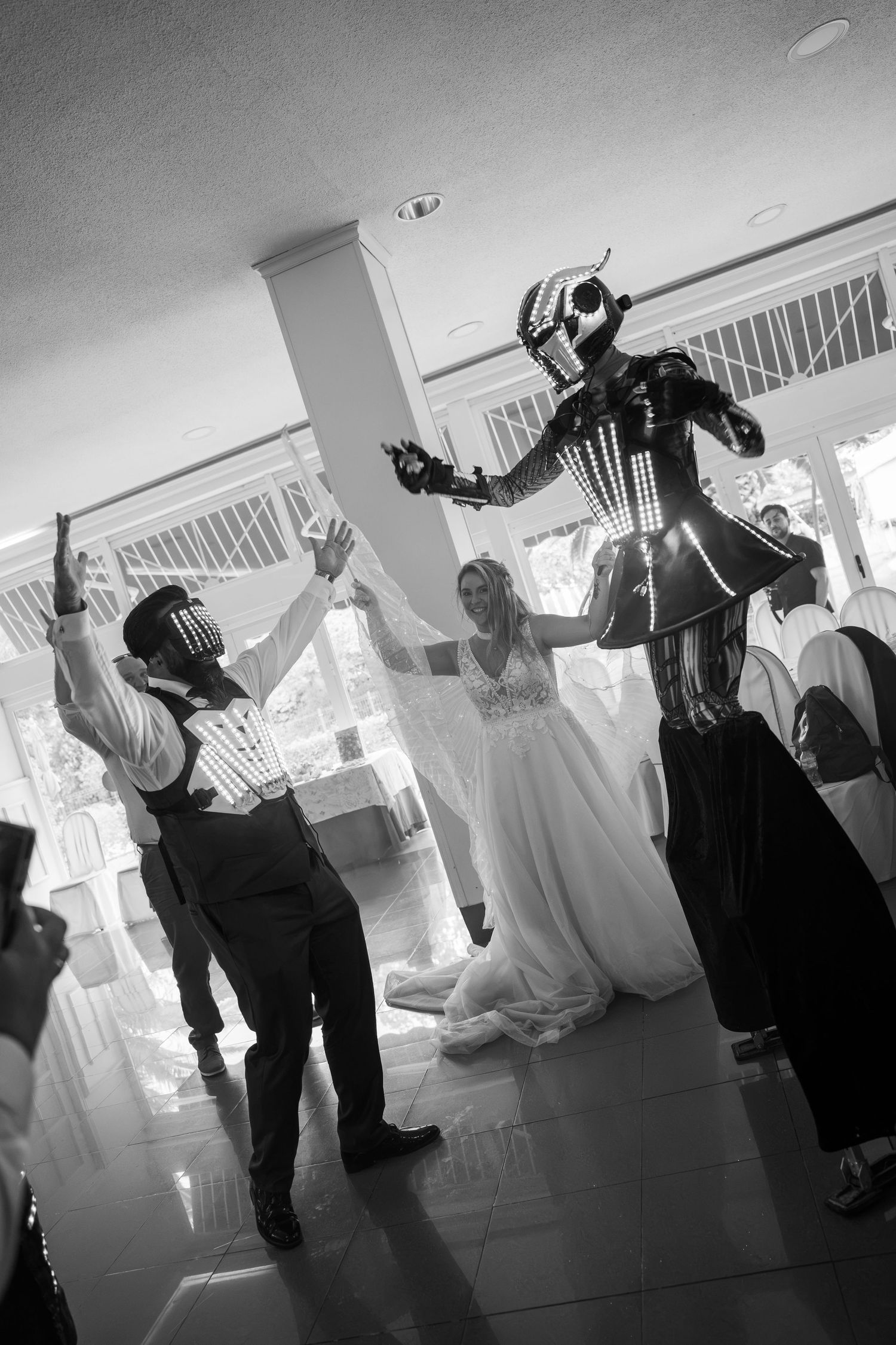 Black and white wedding photo featuring a bride, groom, and LED stilt walker performer celebrating at a reception.