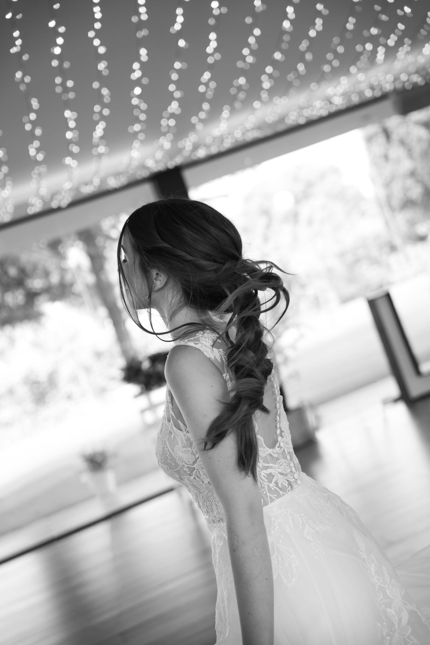 Black and white photo of bride with braided hair in lace wedding dress dancing under twinkling fairy lights.
