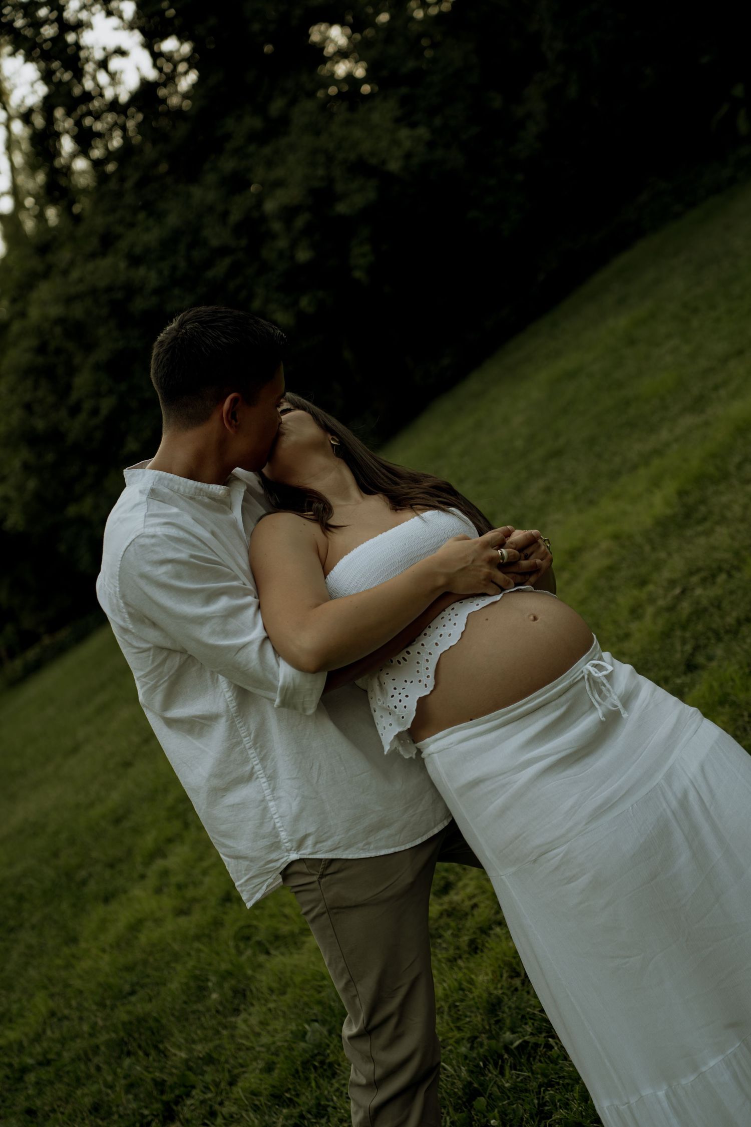 Expecting couple sharing a romantic kiss on a grassy hillside during a maternity photoshoot in white outfits.
