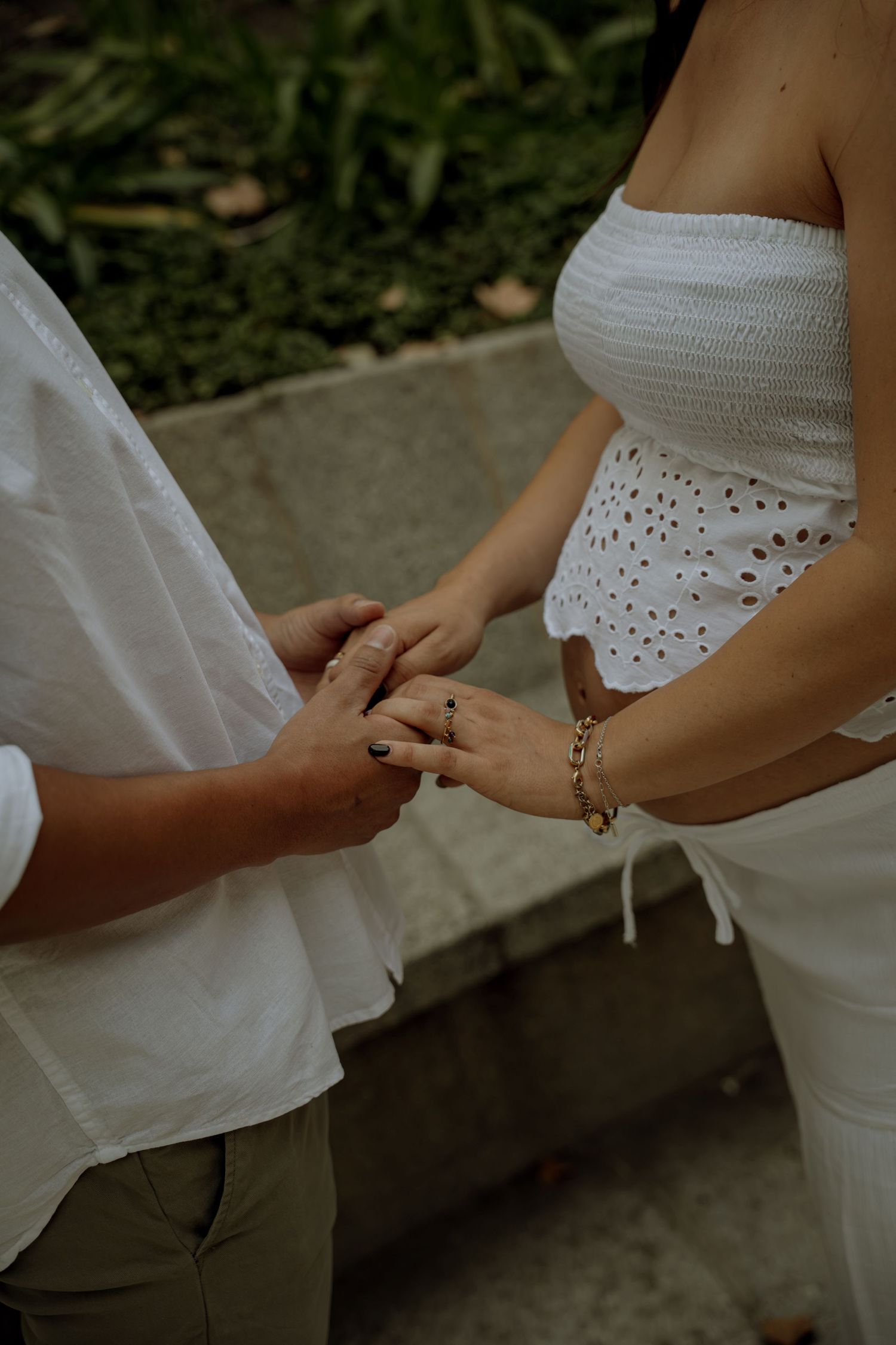 Close-up of couple holding hands, pregnant woman in white eyelet top wearing gold bracelet outdoors.
