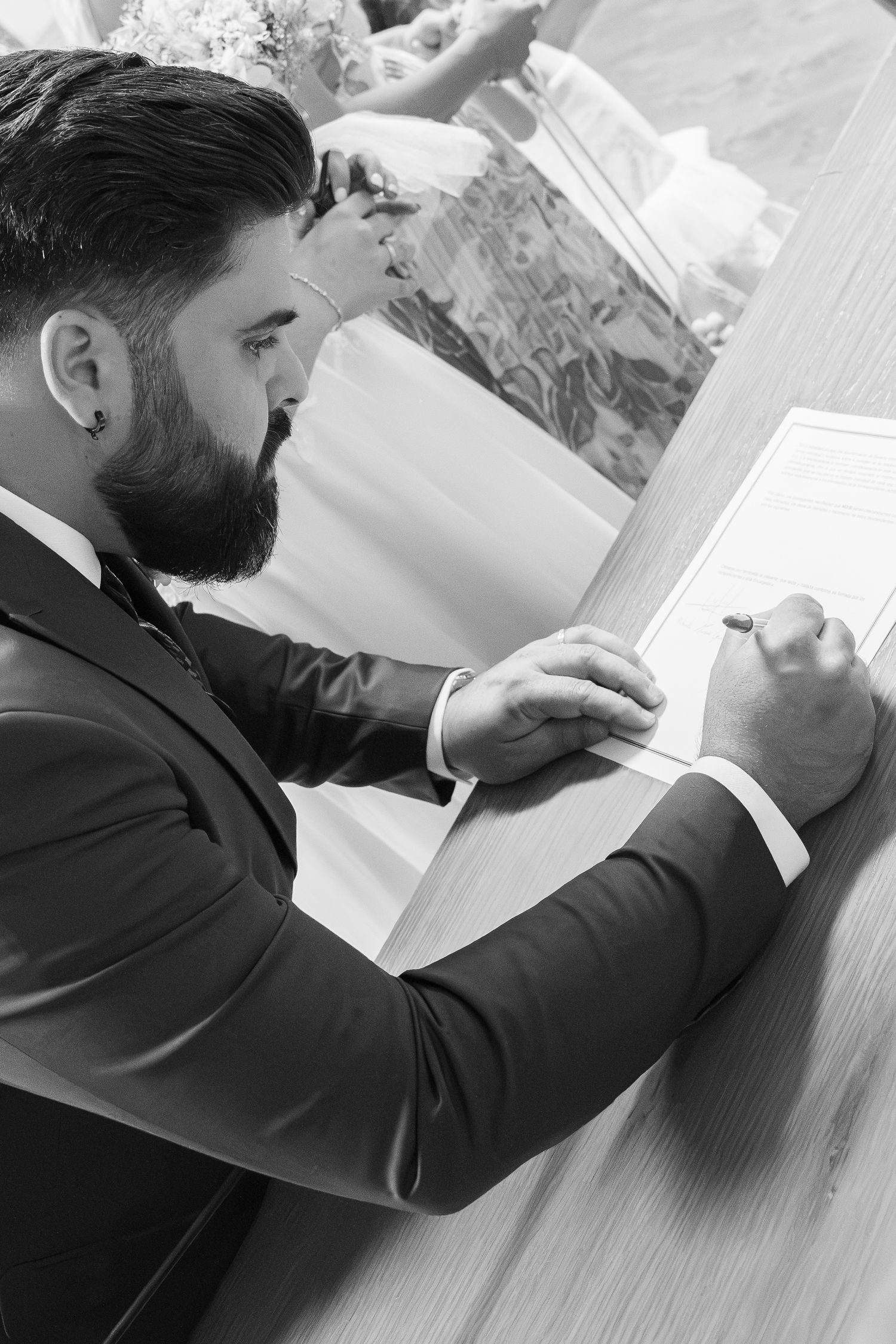 Bearded groom in a dark suit and tie signs the marriage register at a wooden table while a bride in a white gown sits beside him.