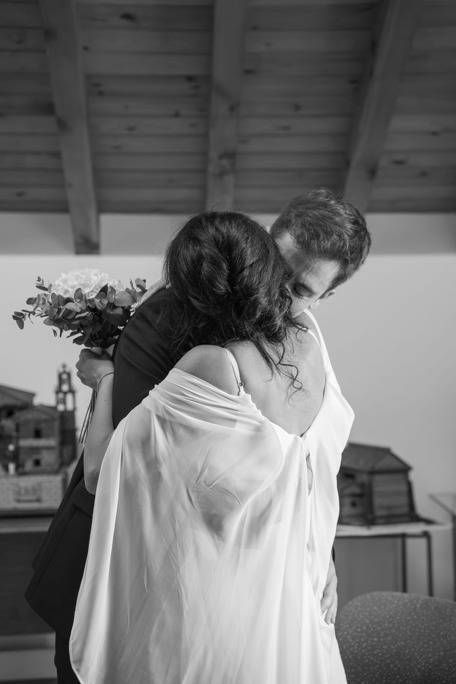 Black and white photo of a bride in a sheer white wrap embracing a groom while holding a bouquet in a rustic room with wooden beams.