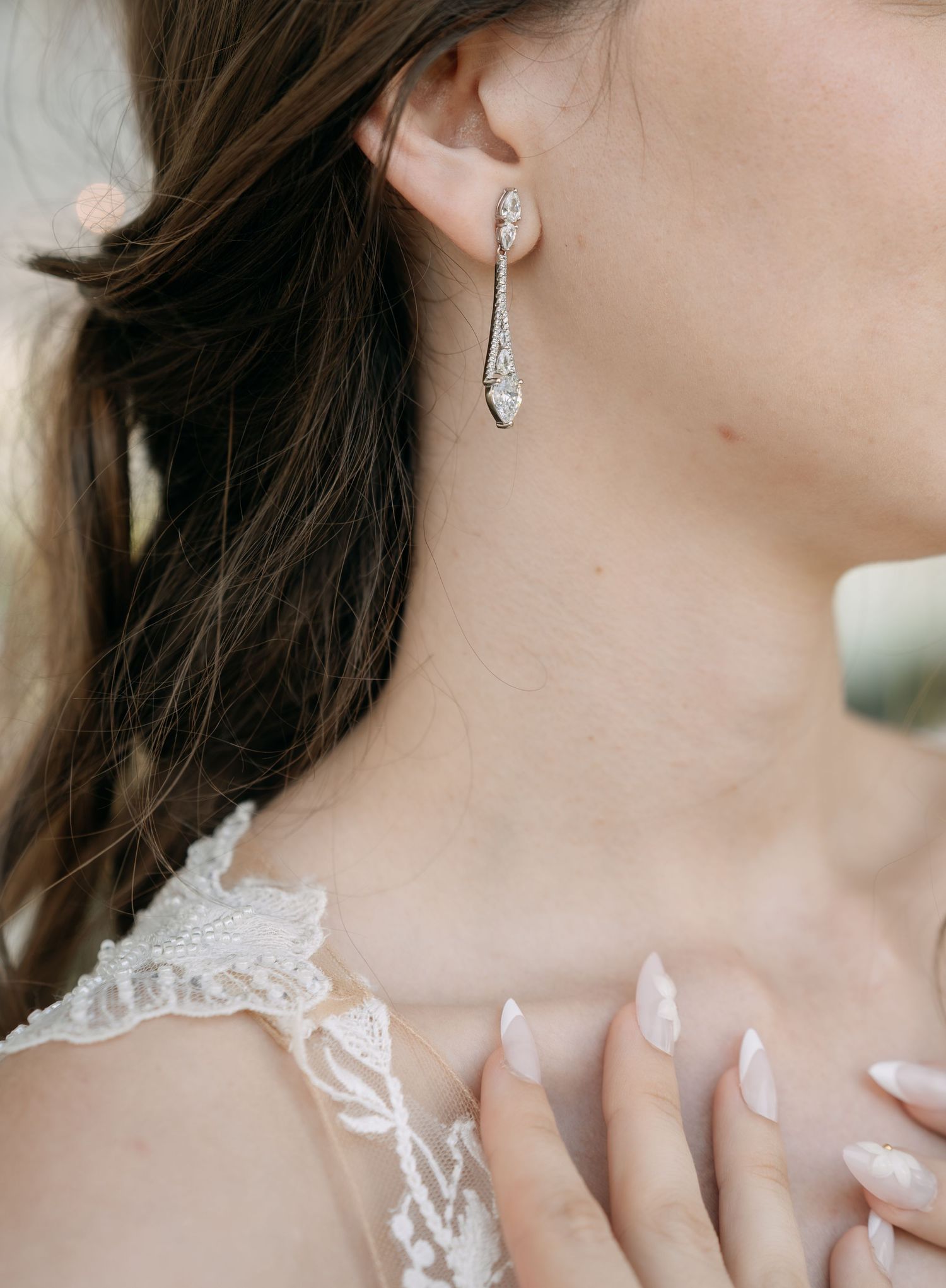 Bride wearing elegant silver crystal drop earrings and a lace gown with her hand resting on her chest, showing a soft manicure.