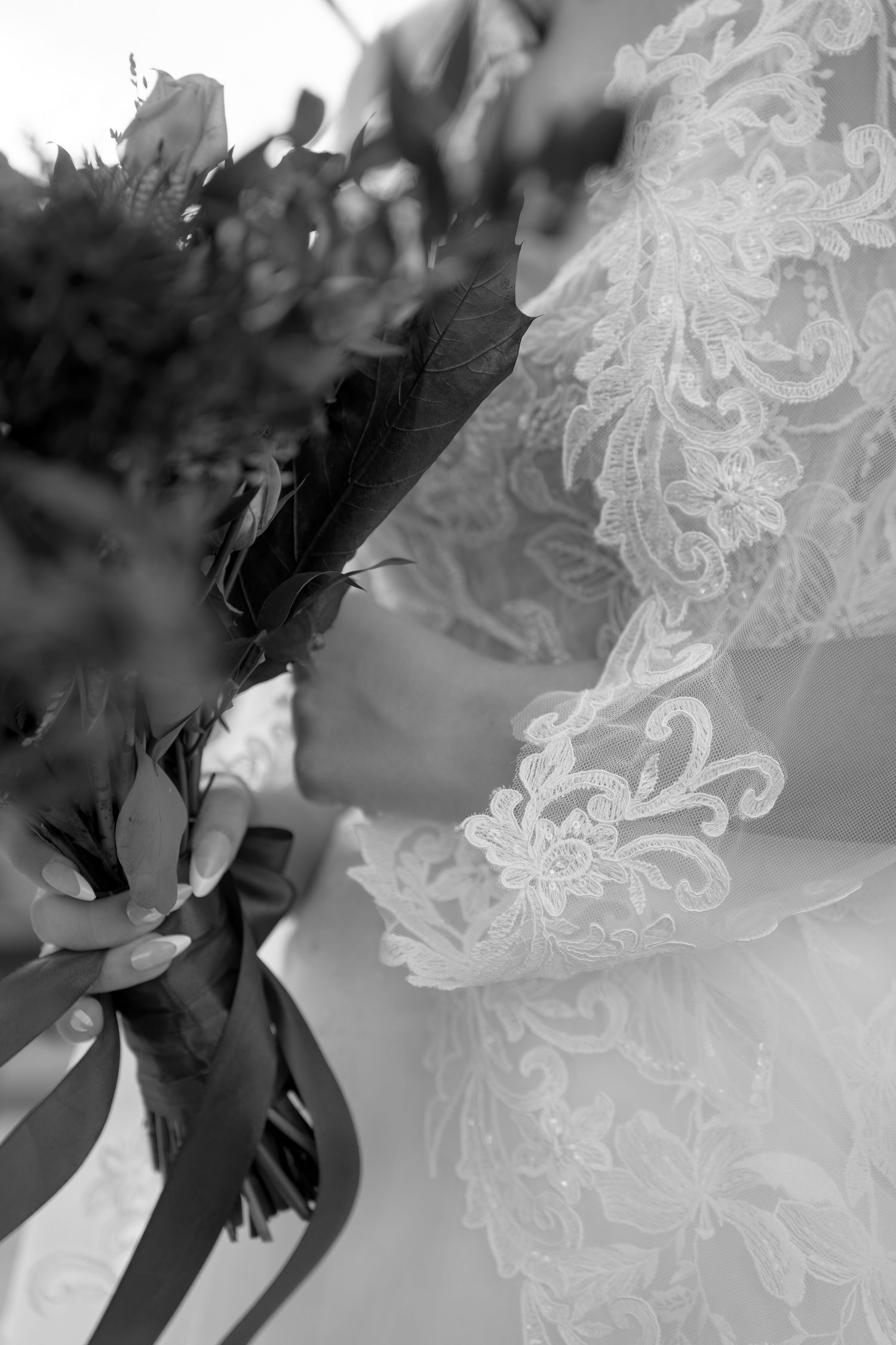Black and white close-up of a bride in a lace wedding dress with long sleeves holding a bouquet tied with satin ribbons.
