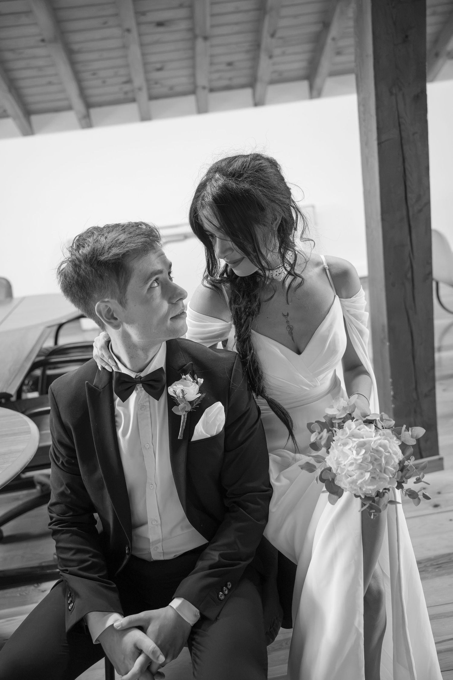 Black and white photo of a bride in a white gown holding a bouquet, gazing down at a seated groom in a suit and bow tie.