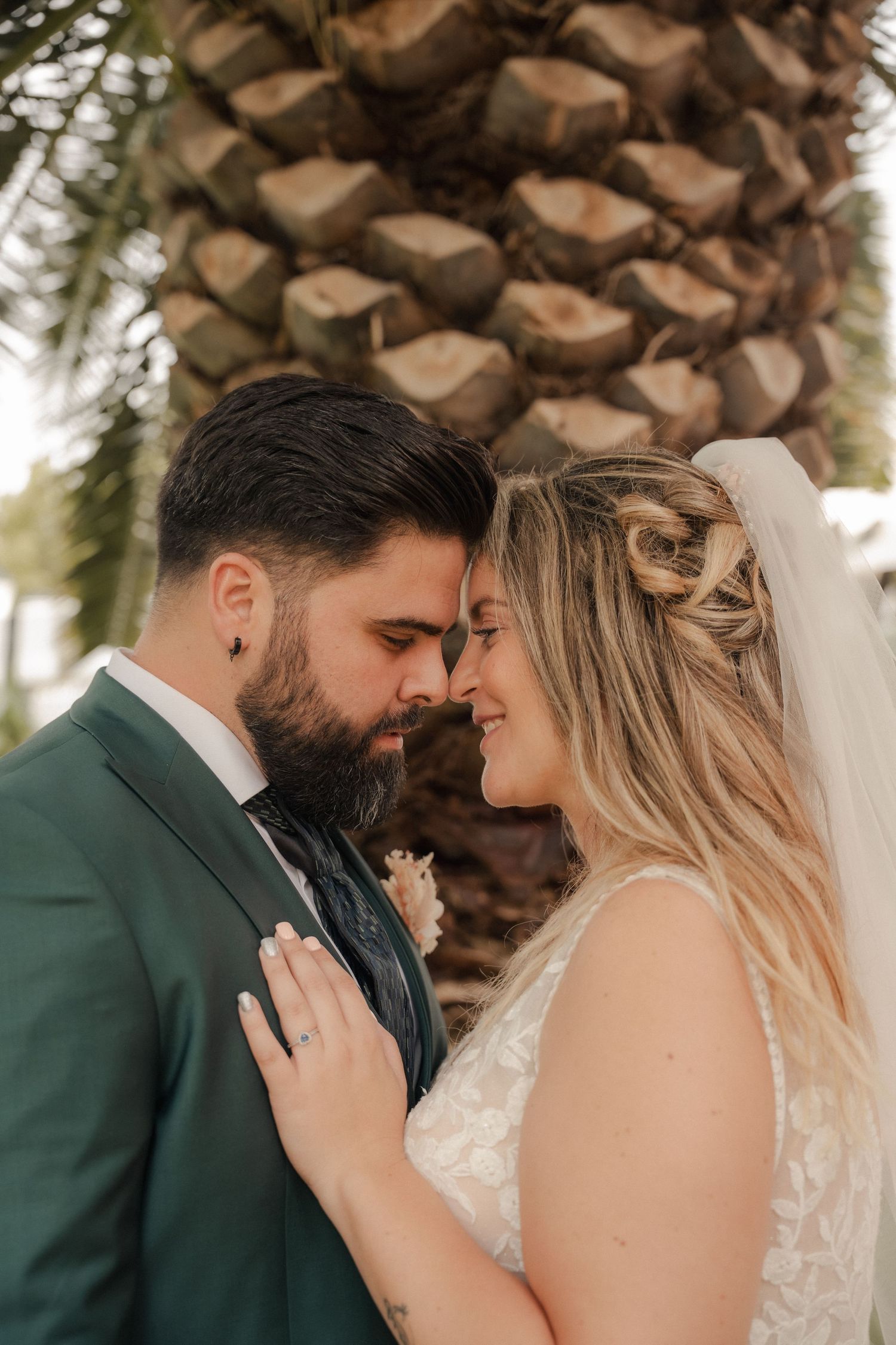 Bride in a lace wedding dress and veil touches foreheads with her bearded groom in a green suit in front of a palm tree.