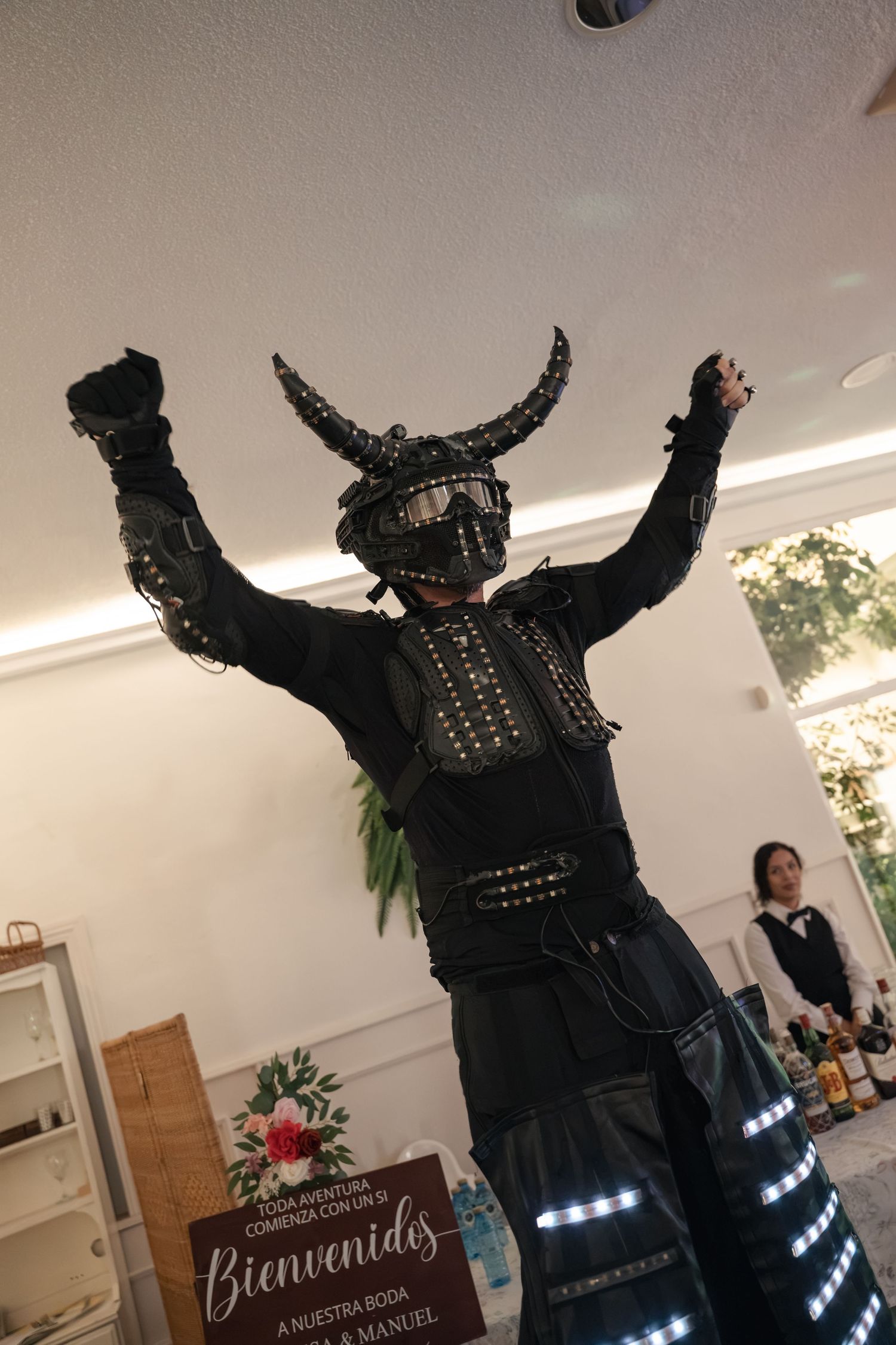 Performer in a black horned helmet and LED-lit armor costume raises their arms at an indoor event, with a welcome sign and server behind.