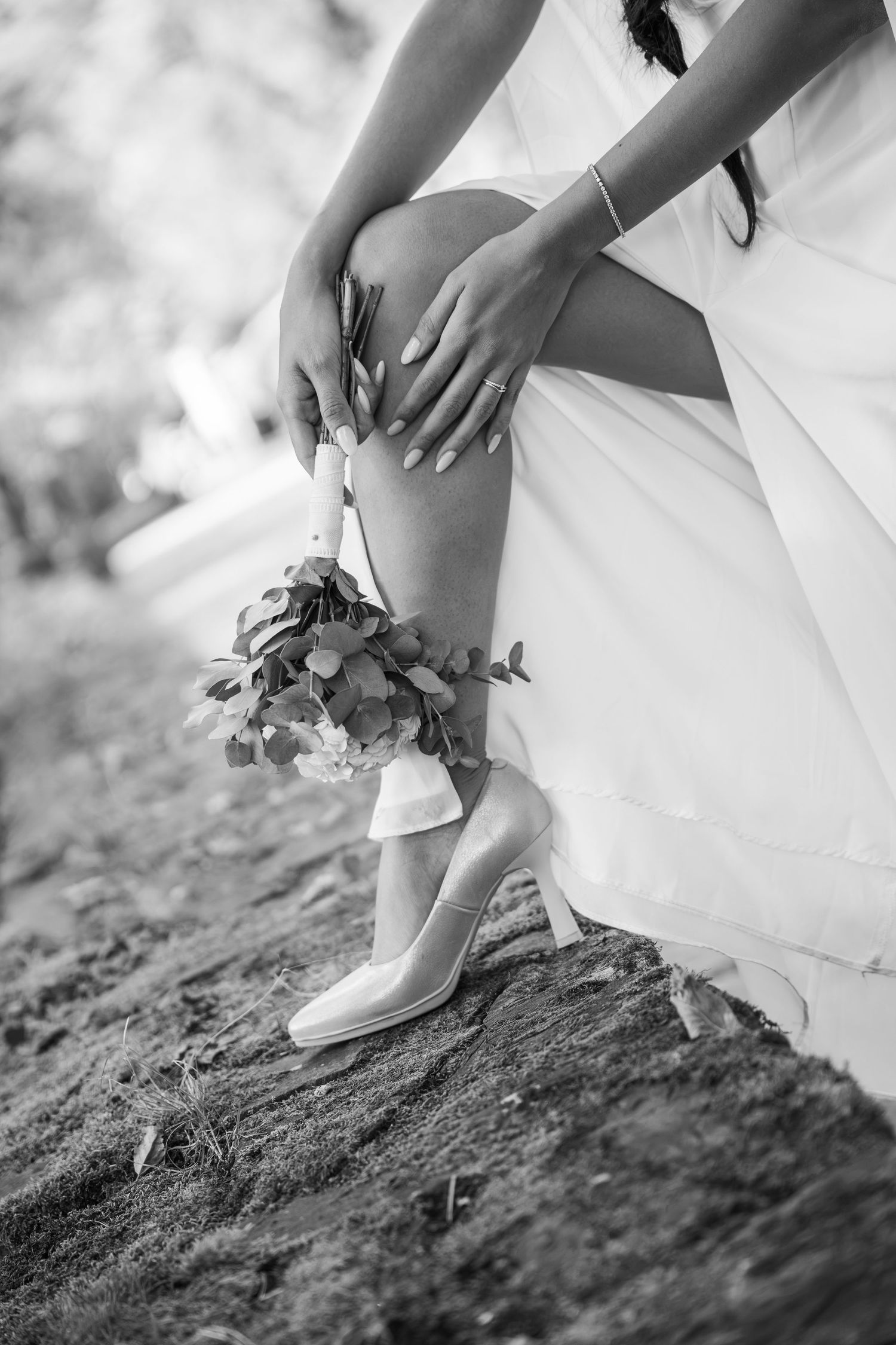 Black and white photo of a bride in a flowing gown and heels, kneeling on rocky ground while holding a leafy wedding bouquet.
