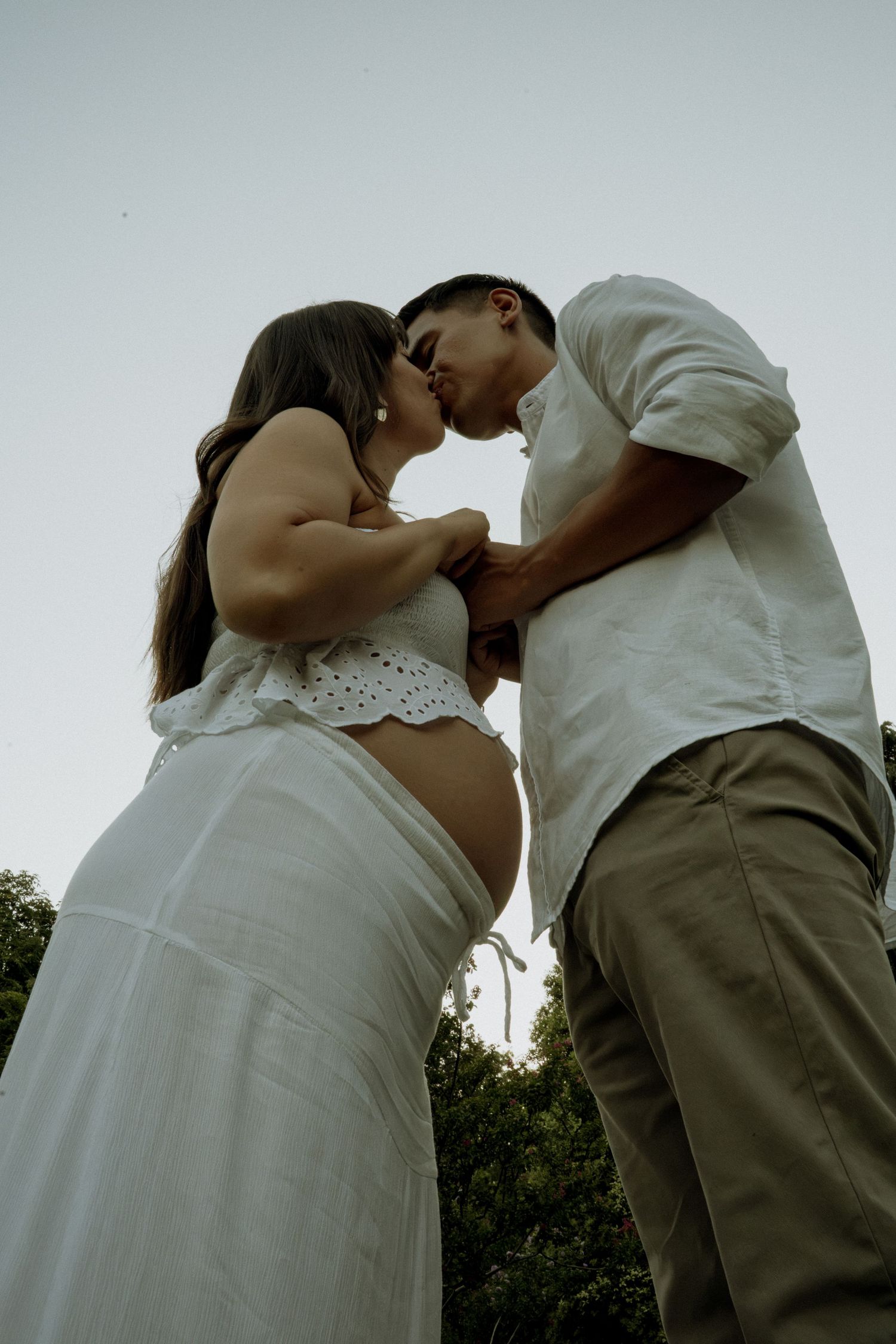 Low-angle view of a pregnant couple sharing a kiss outdoors at dusk, both dressed in white and beige outfits.