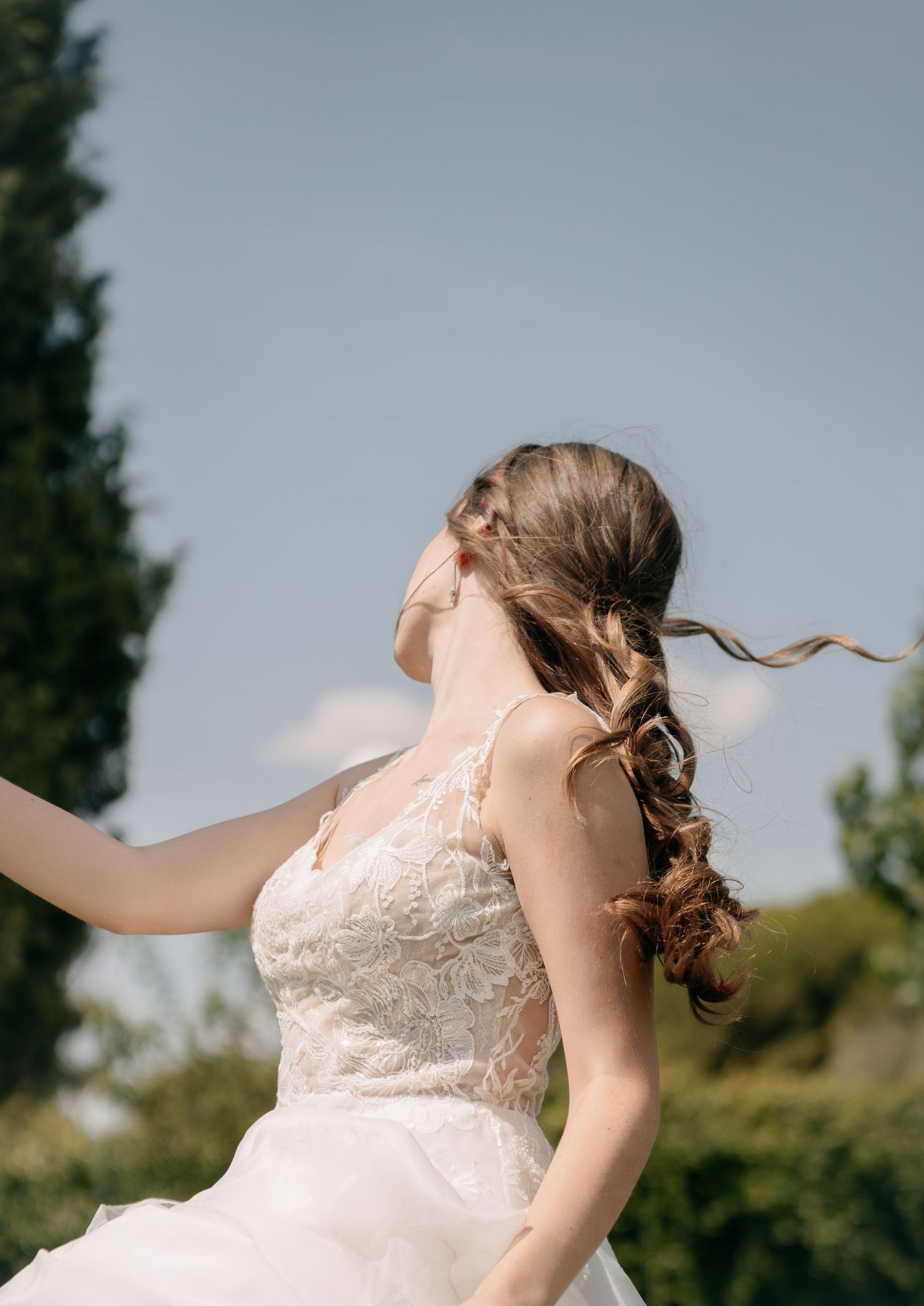 Bride in lace wedding dress with braided hair spinning outdoors against blue sky and green trees.