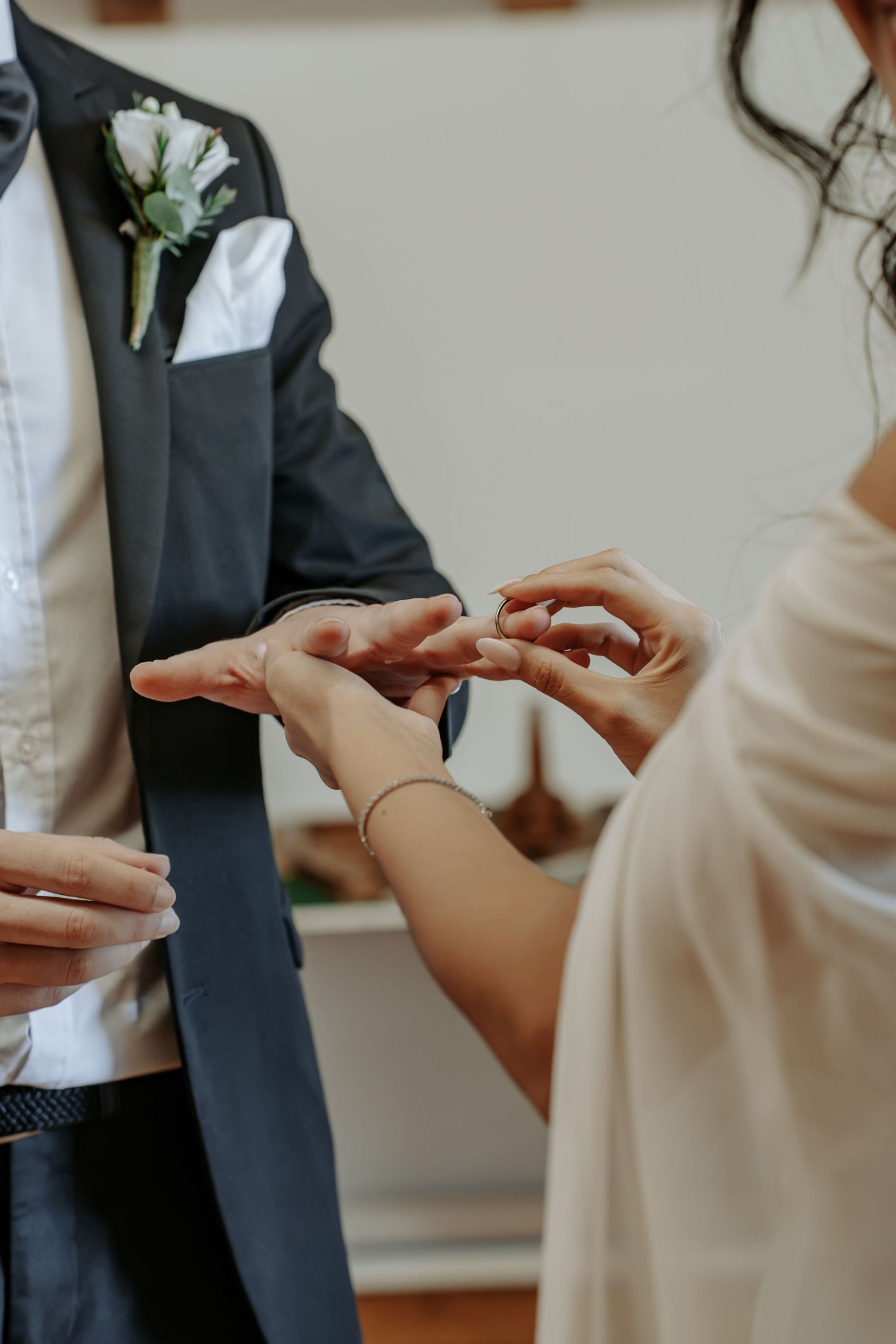 Bride placing a wedding ring on groom's finger during ceremony, groom wearing navy suit with floral boutonniere.