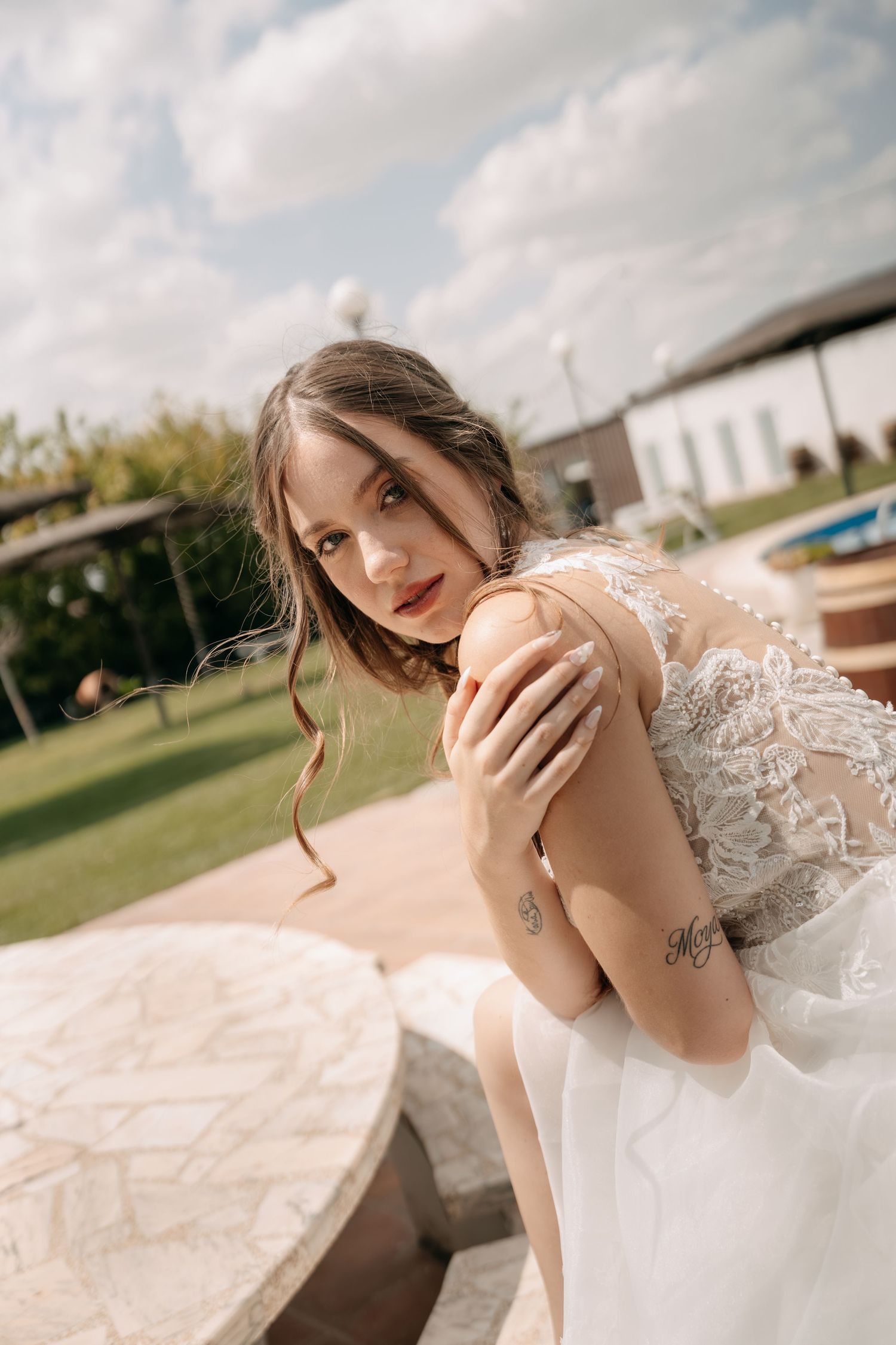 Bride in a lace wedding gown with loose wavy hair poses on a stone terrace, glancing over her shoulder with a lawn and cloudy sky behind.