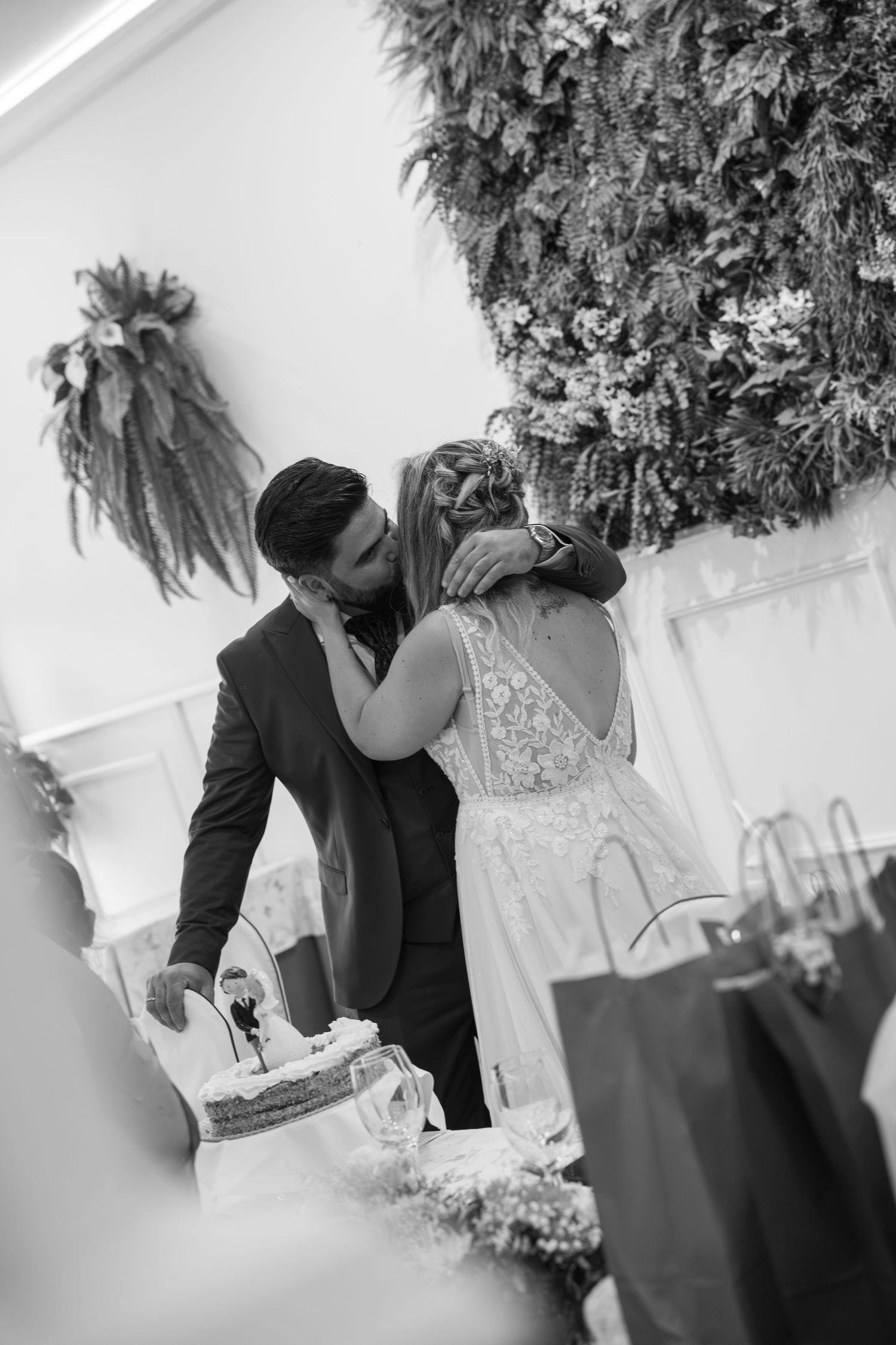 Black and white photo of a bride and groom kissing beside a wedding cake, with a lush hanging floral wall behind them.