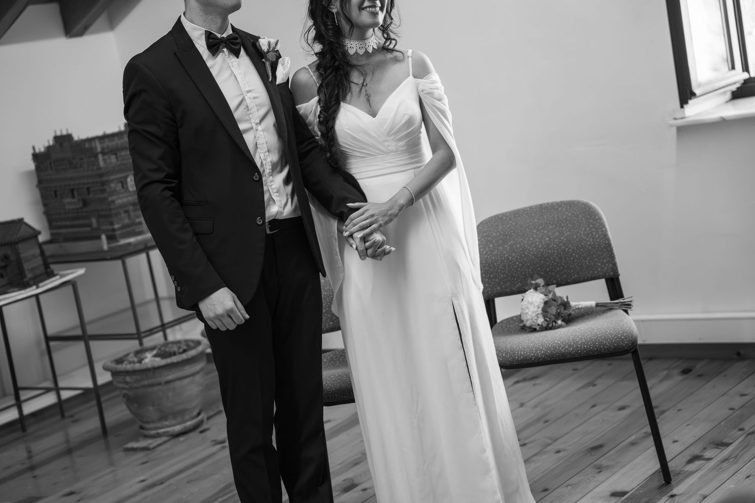 Black and white photo of a bride in a flowing gown and a groom in a suit and bow tie holding hands during an intimate wedding ceremony.