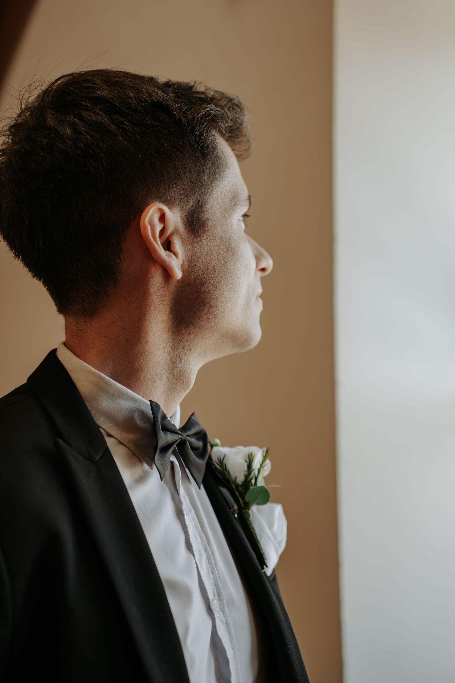 Groom in a black suit, gray bow tie and white floral boutonniere gazes toward a window in soft natural light before the wedding.