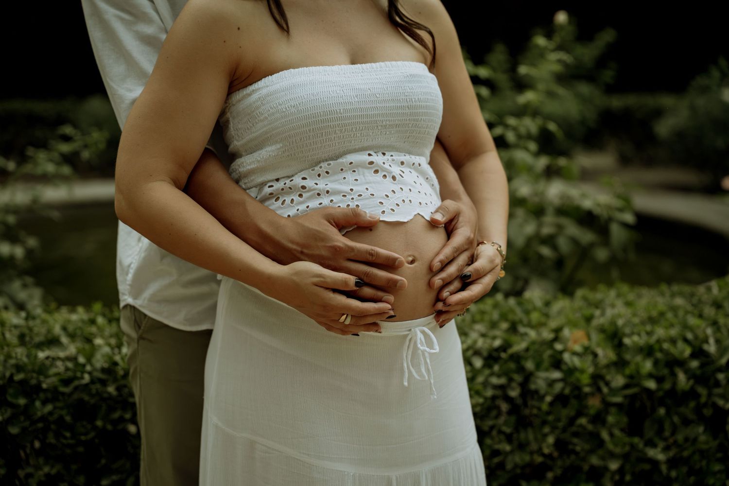 Close-up of couple's hands cradling pregnant belly, woman wearing white lace maternity outfit outdoors.
