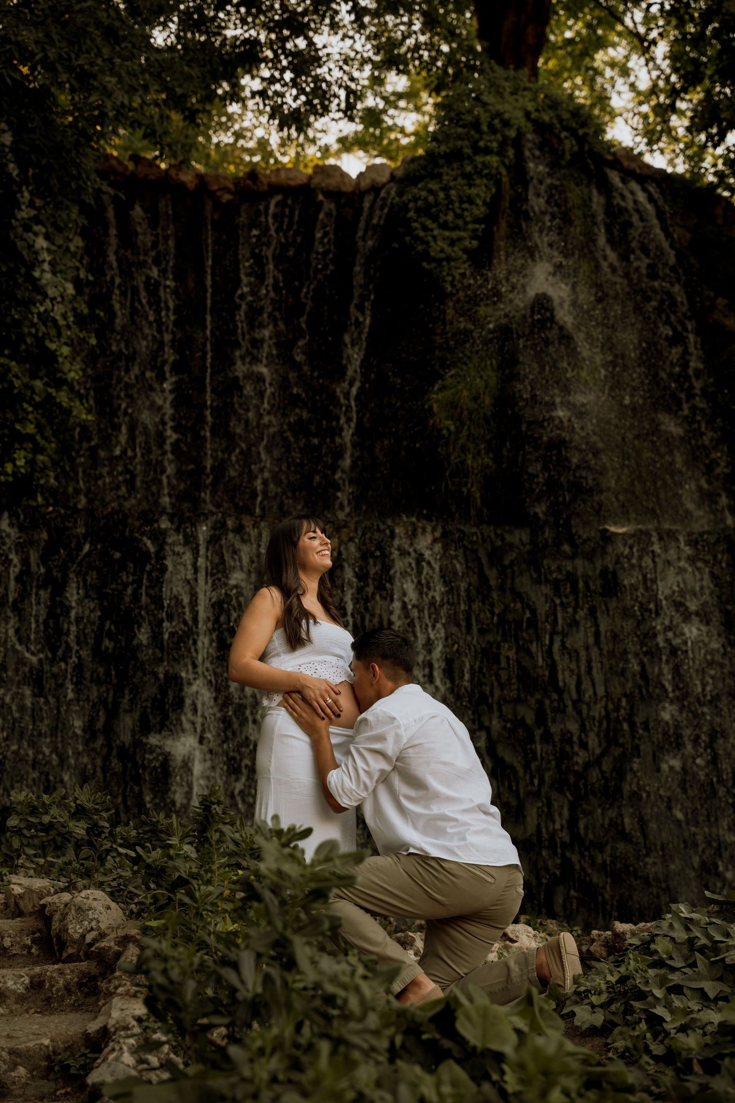 Man kneeling to embrace pregnant woman's belly near a mossy waterfall wall during an outdoor maternity session.