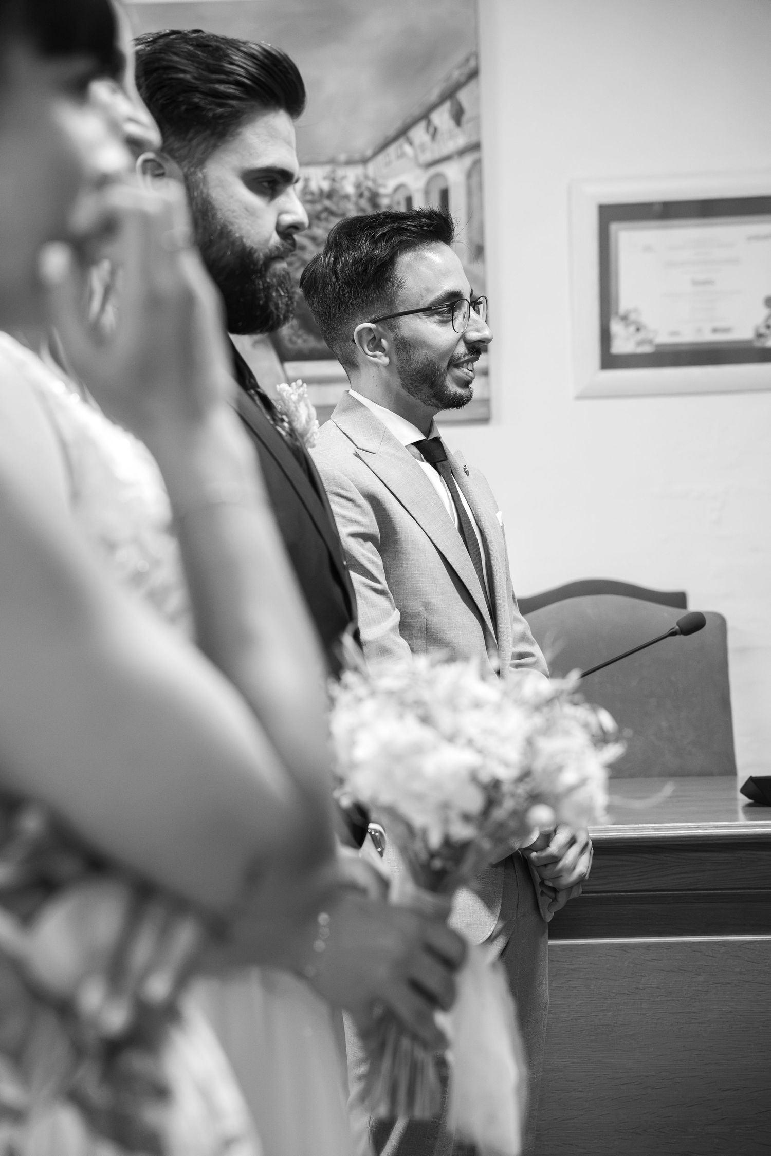Black and white photo of two smiling men in suits at a civil wedding ceremony, with a blurred bride holding a bouquet in the foreground.