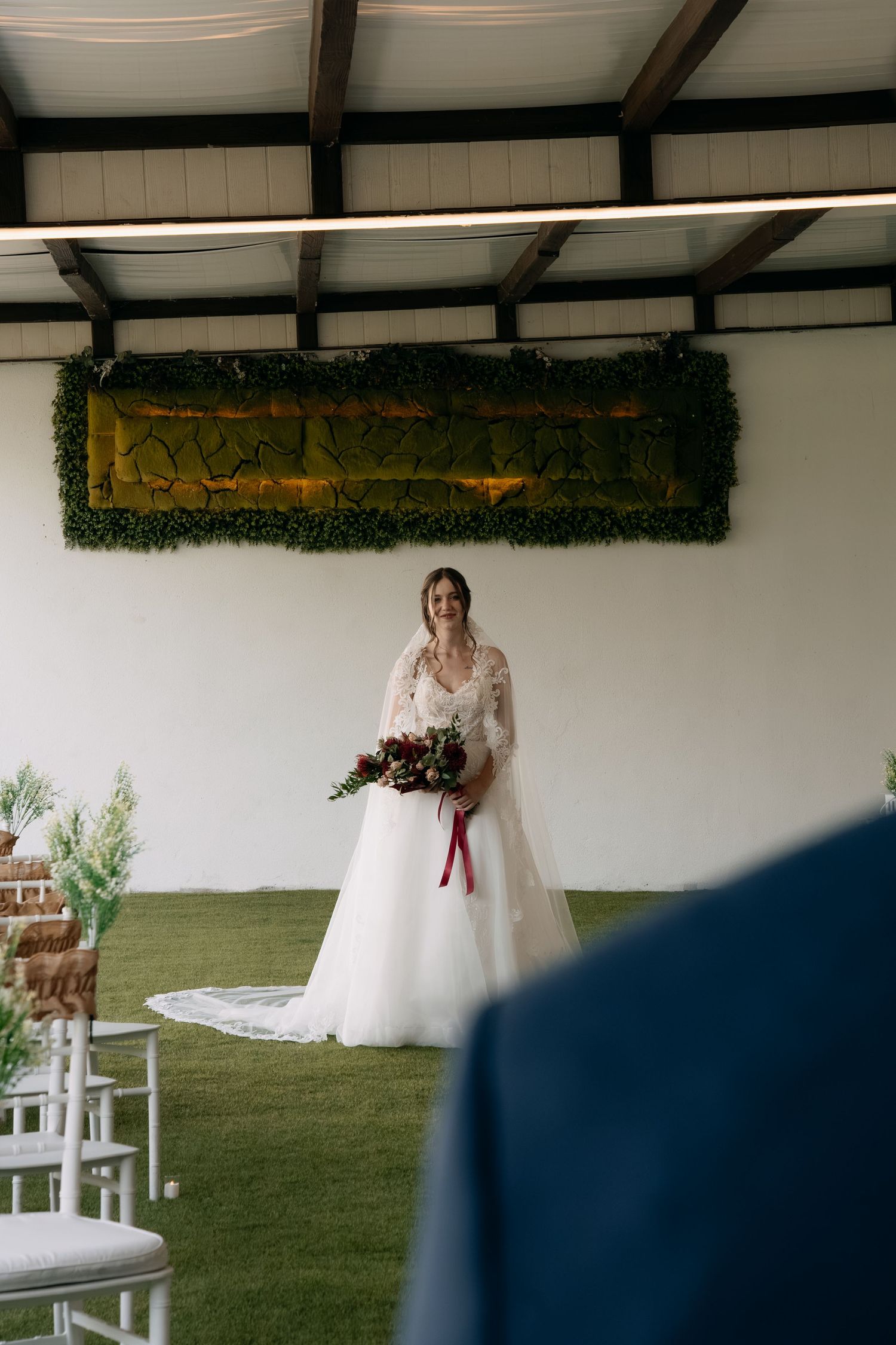 Bride in a white lace gown and long veil holds a burgundy bouquet while standing on green grass at an indoor wedding ceremony venue.