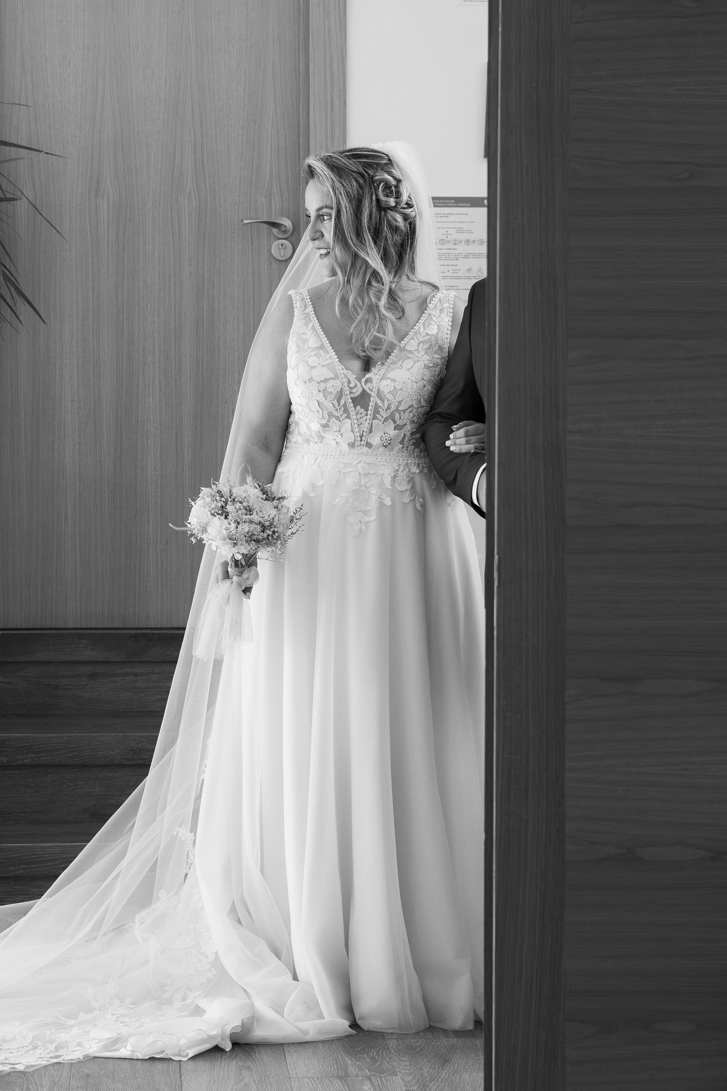 Black and white photo of a bride in a lace wedding gown holding a bouquet, standing in a doorway with her groom.