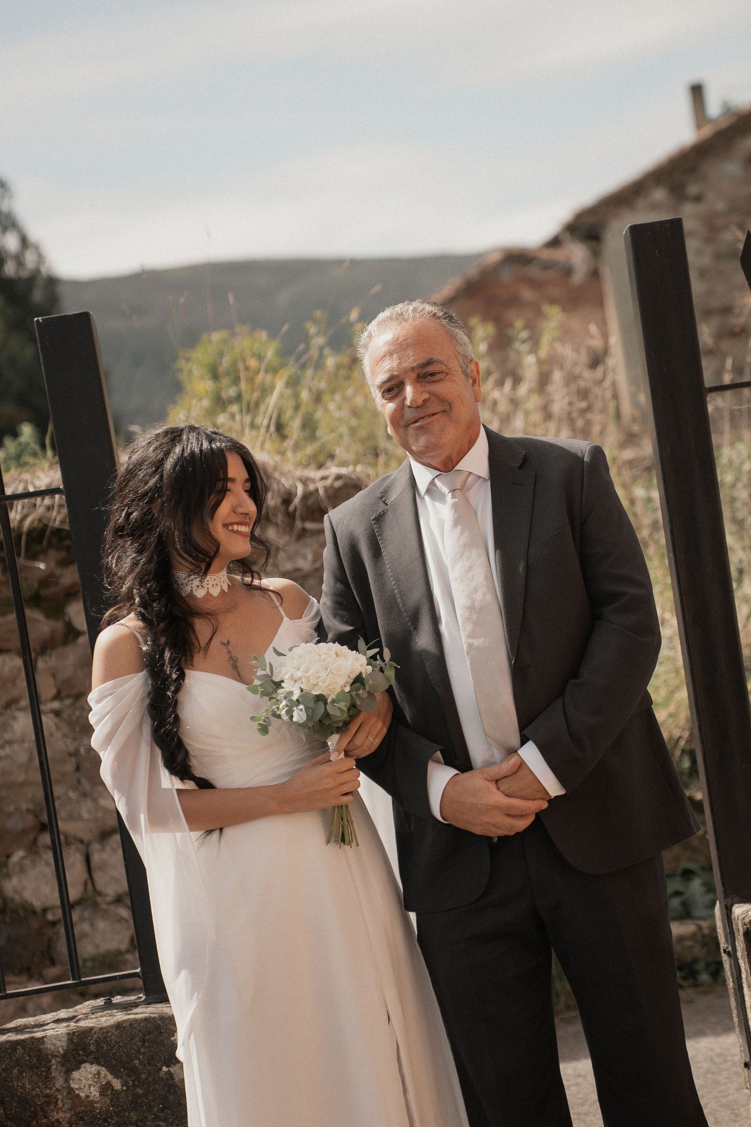 Smiling bride in a flowing white dress holding a white bouquet stands beside a man in a dark suit at a rustic outdoor wedding.