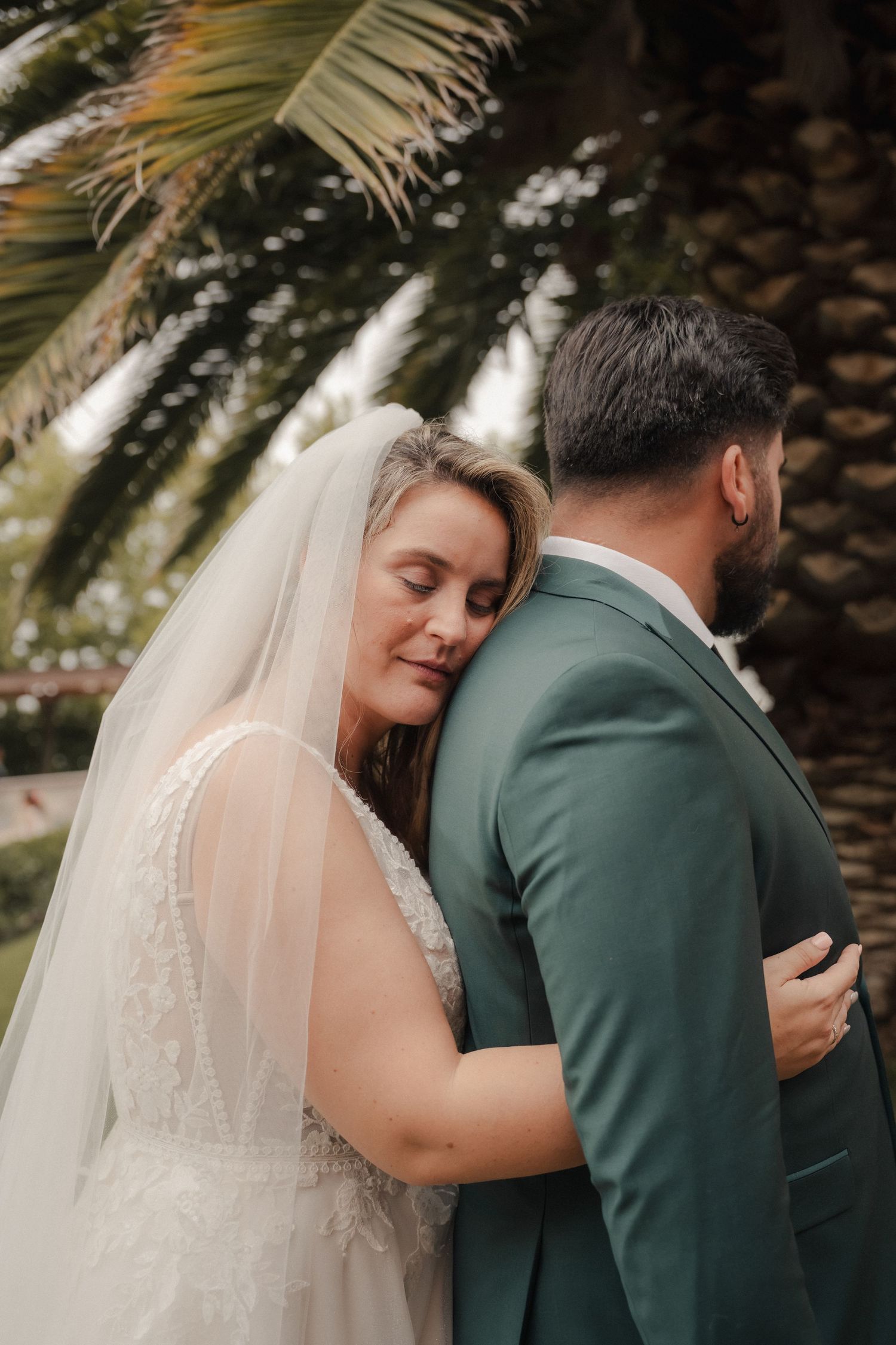 Bride in a lace gown and long veil rests her head on the shoulder of a groom in a green suit beneath palm trees.