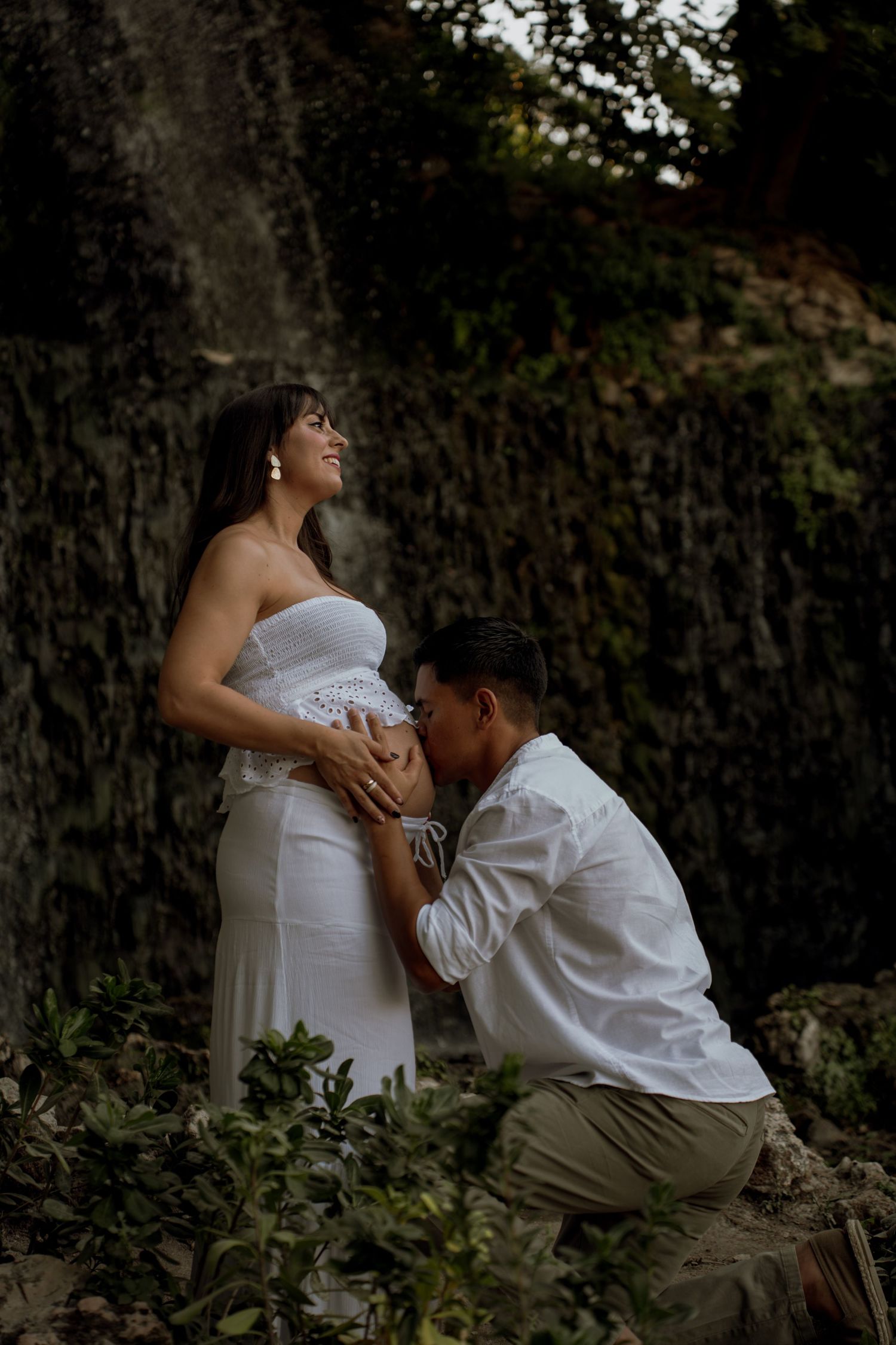 Expectant couple in white outfits share tender moment outdoors, man kneeling kissing pregnant belly near large tree.