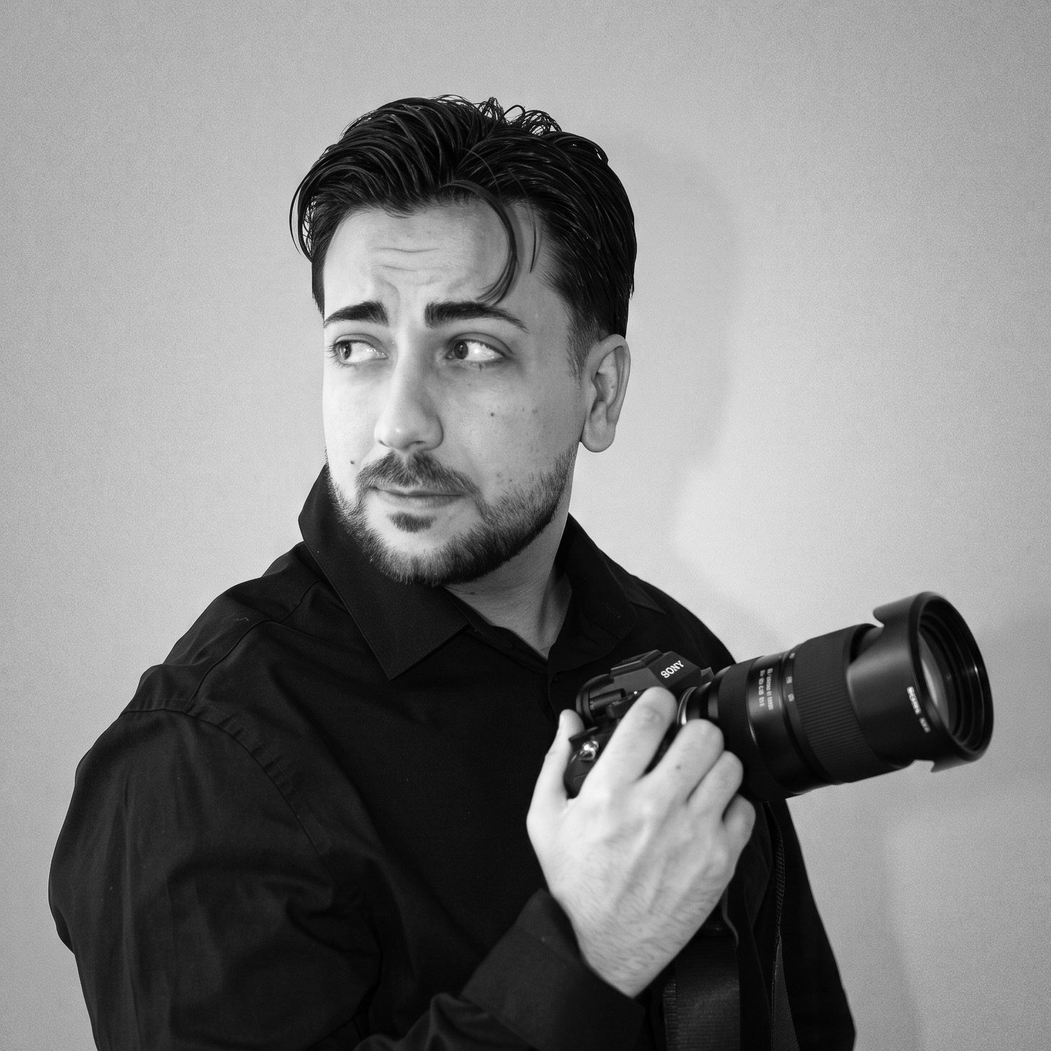 Black and white portrait of a bearded male photographer holding a camera, looking sideways thoughtfully.