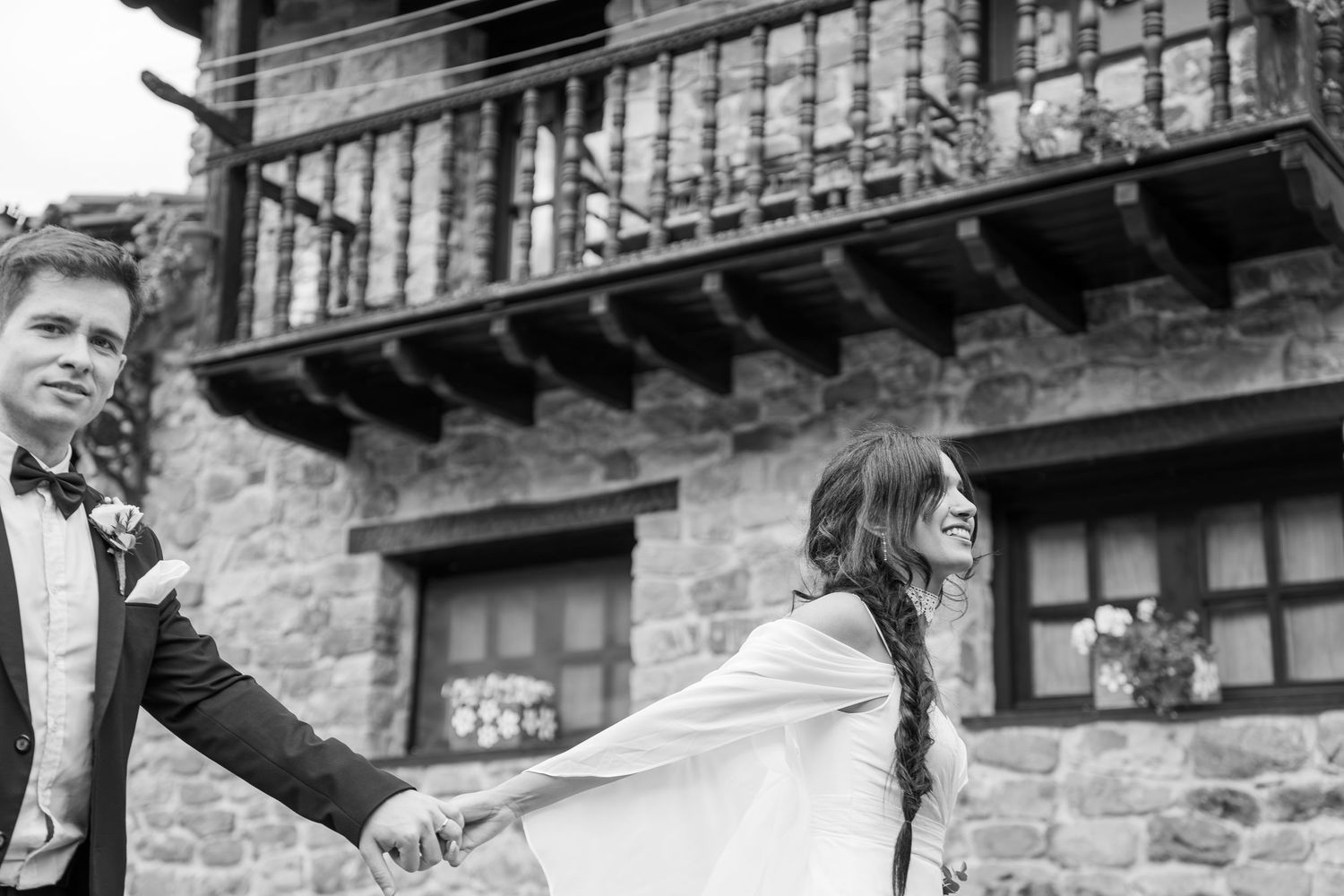 Black and white photo of bride and groom holding hands before a rustic stone building.