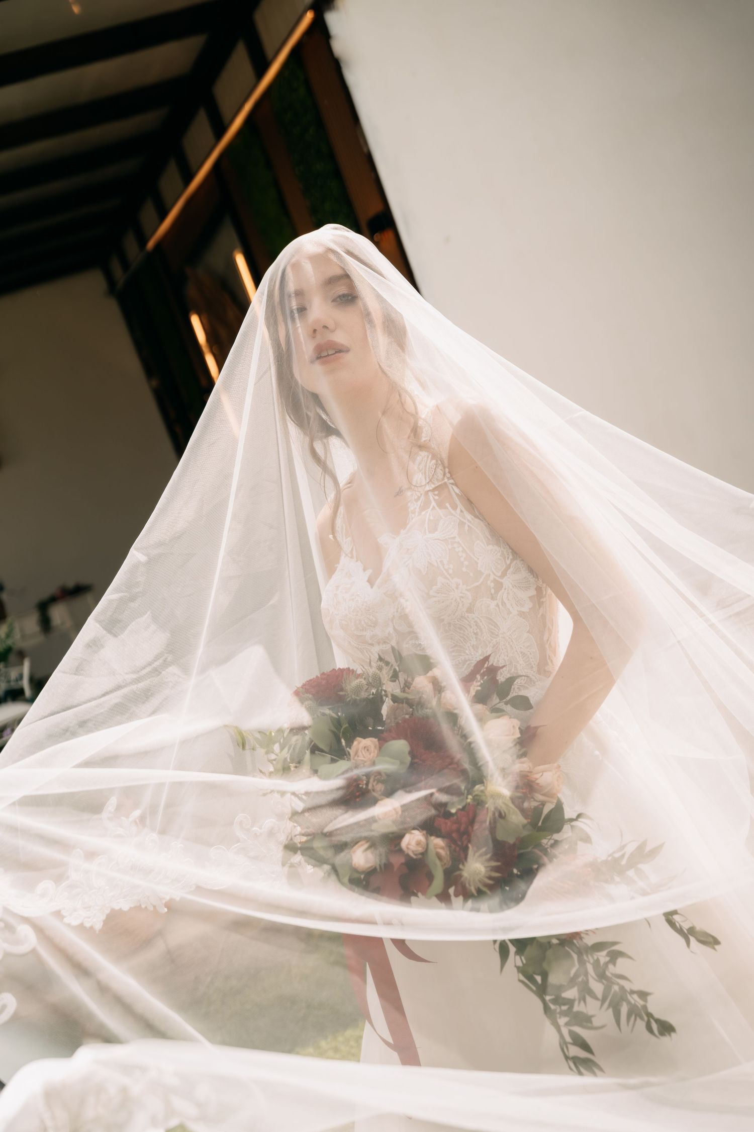 Bride in lace gown holding floral bouquet draped under flowing white veil indoors.