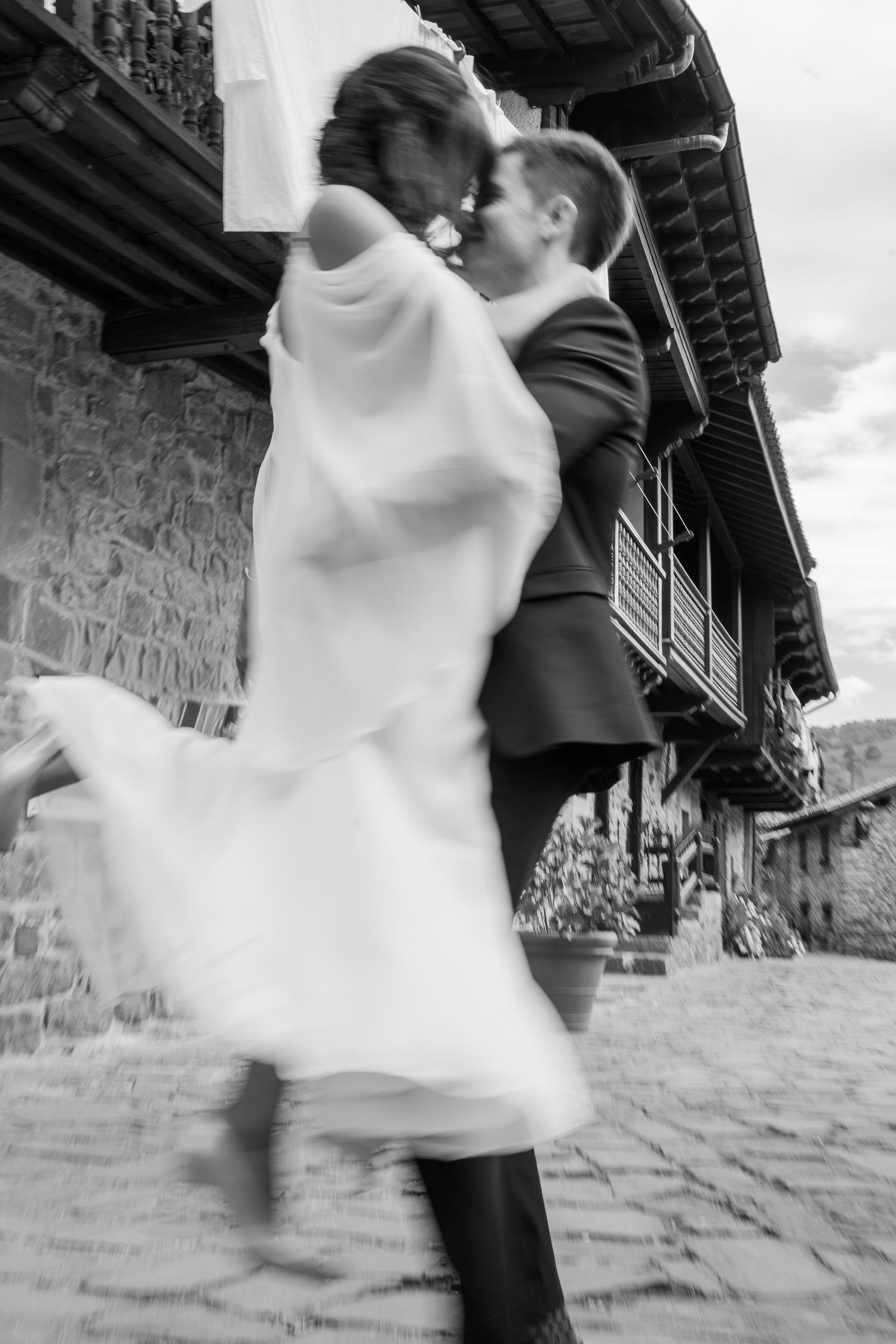 Black and white wedding photo of bride and groom sharing a romantic kiss on cobblestone street near rustic stone building.