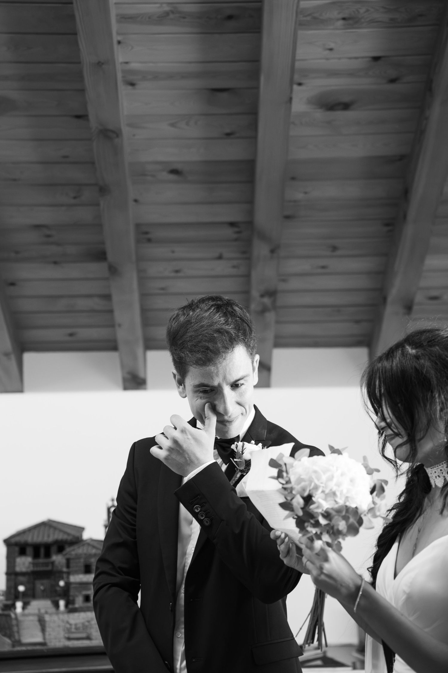 Black and white photo of emotional groom wiping tears beside bride holding bouquet at wedding ceremony.