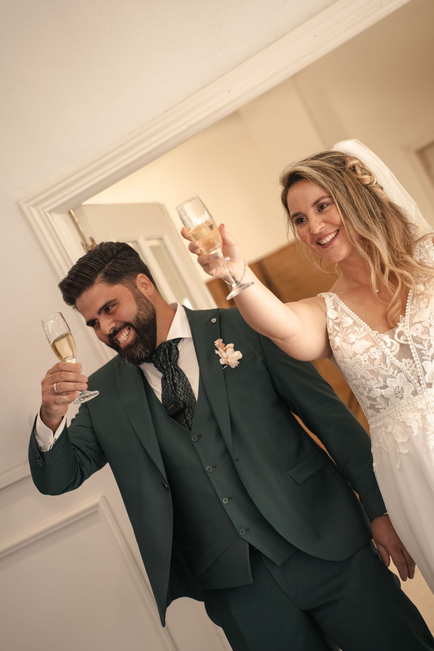 Smiling bride in a lace wedding gown and groom in a green three-piece suit raise champagne glasses in a toast at their wedding.