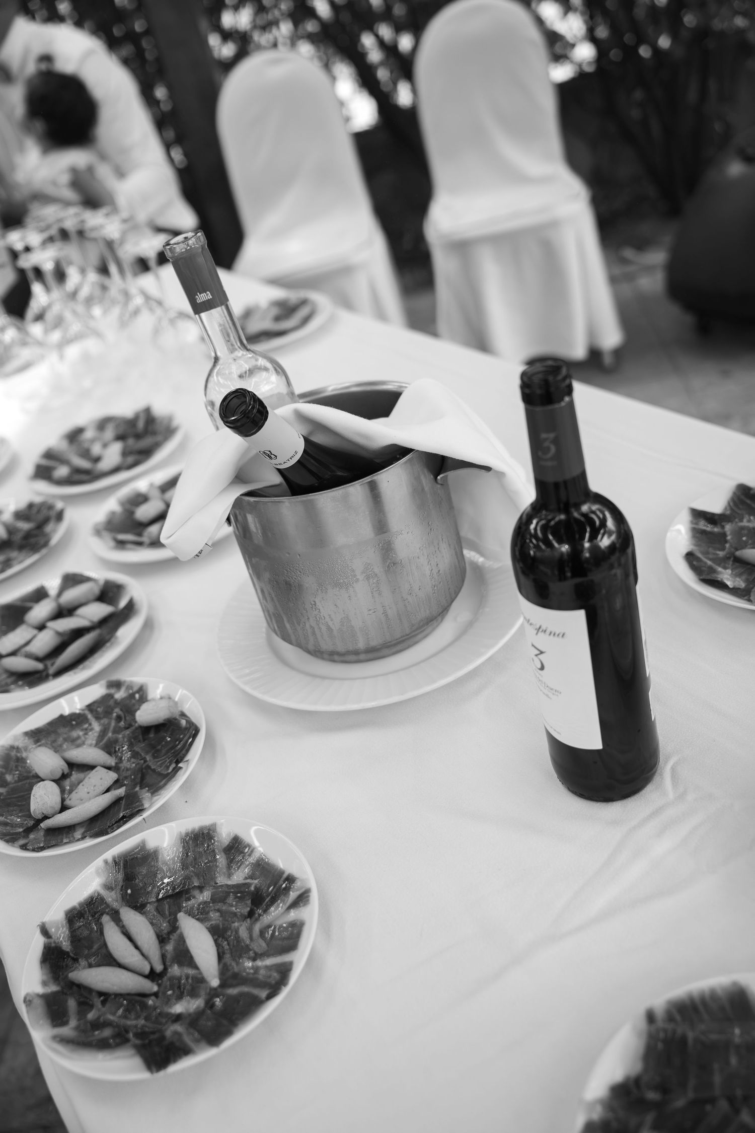 Black and white photo of a banquet table with plates of sliced cured ham, a wine bottle and a bottle chilling in a steel ice bucket.