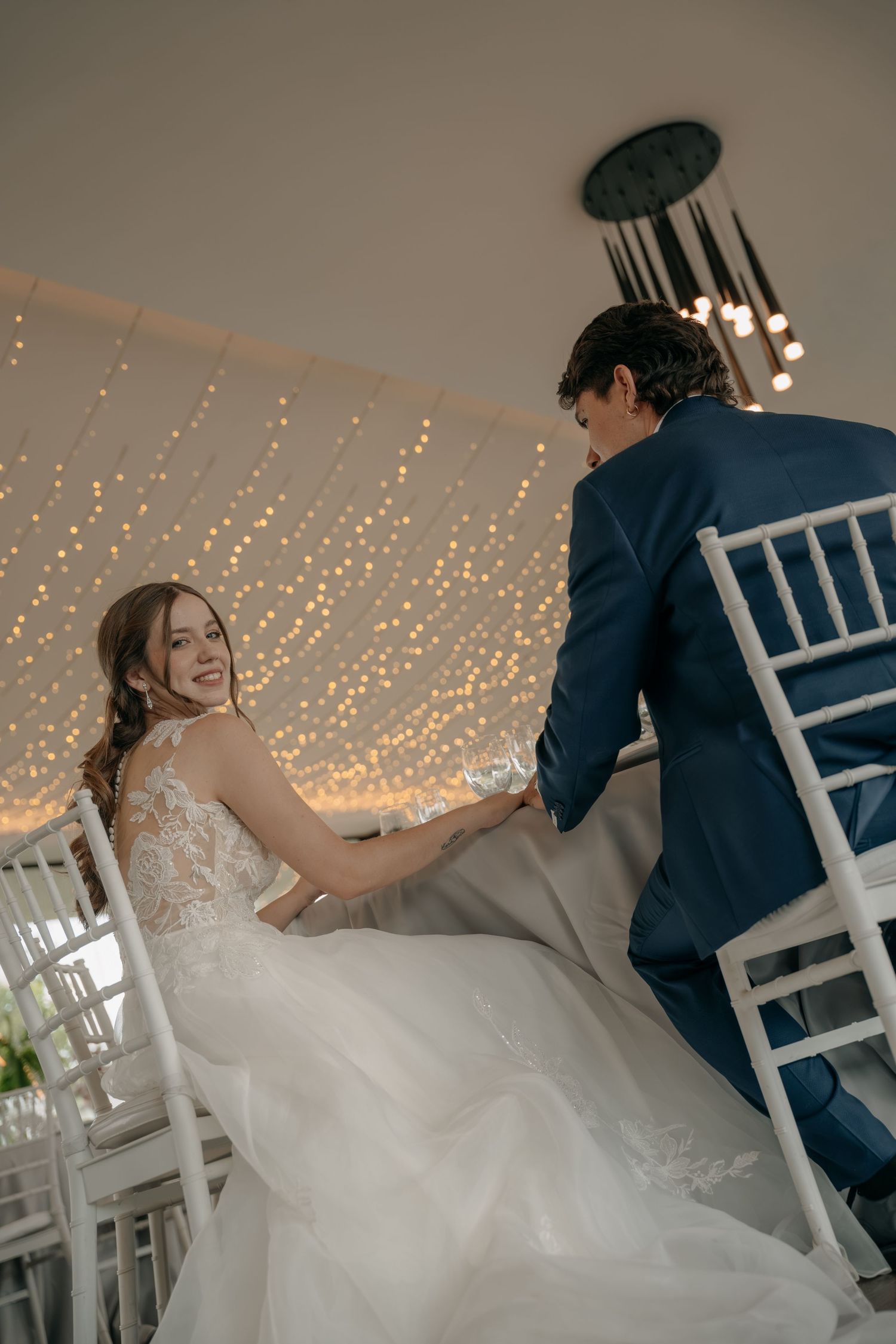 Smiling bride in a lace wedding gown looks back over her shoulder beside a groom in a navy suit under twinkling string lights.