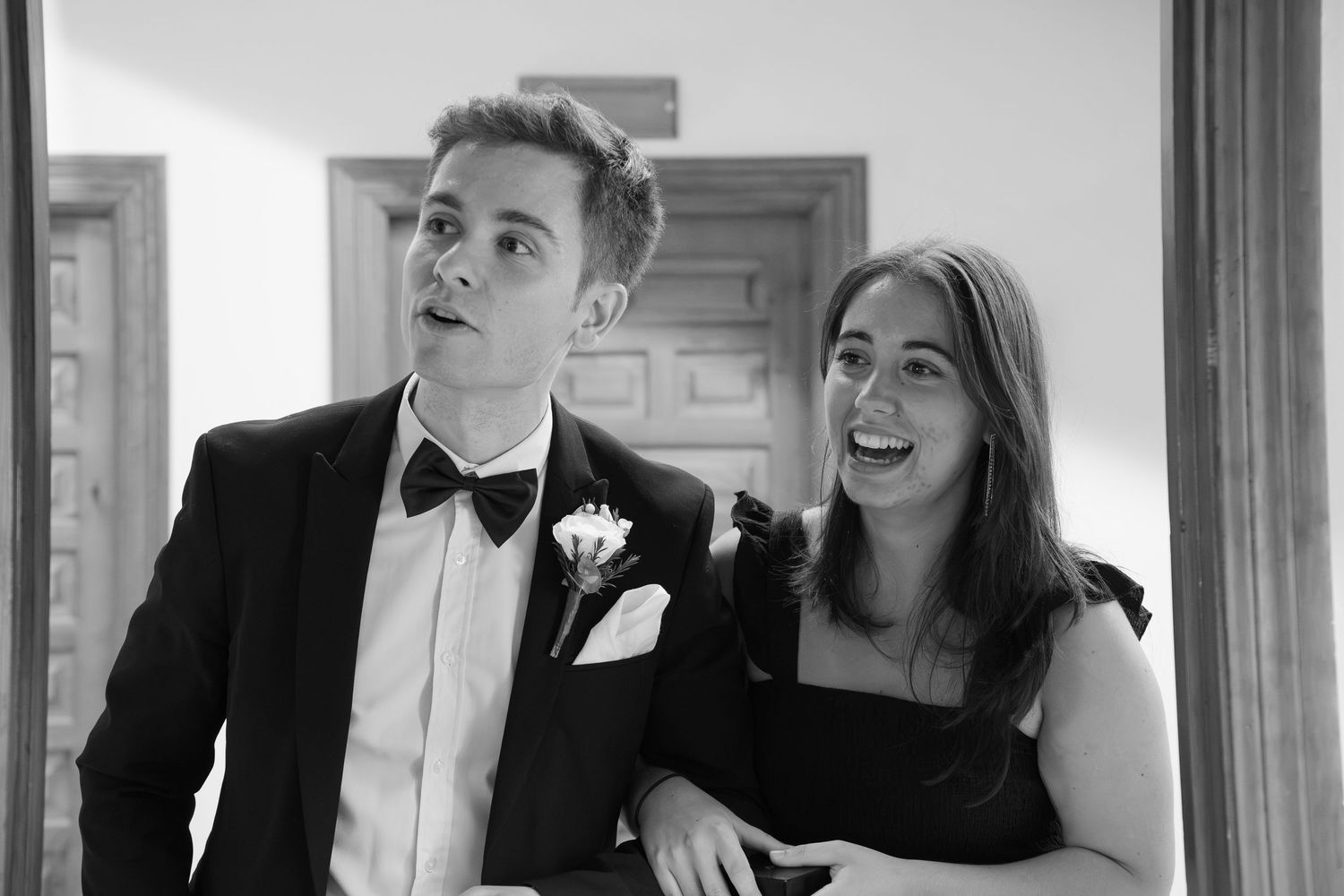 A young man in a tuxedo with a bow tie and boutonniere stands beside a smiling woman in a black dress at a formal event.