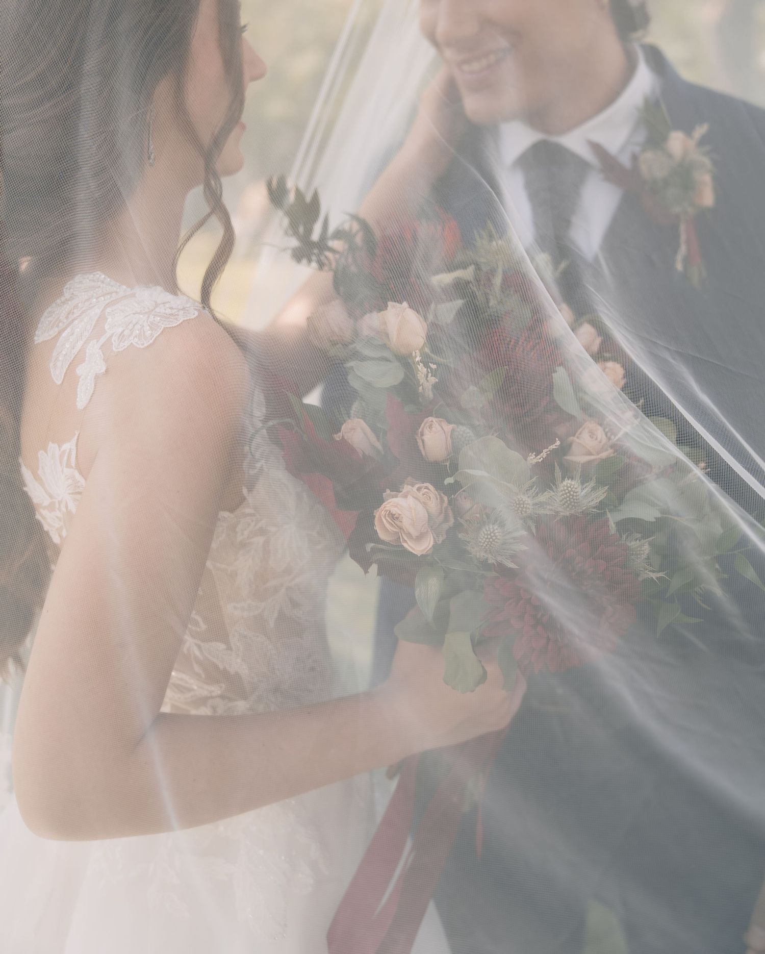 Bride holding a lush bouquet of blush and burgundy roses, standing beside a groom, partially veiled in soft tulle.