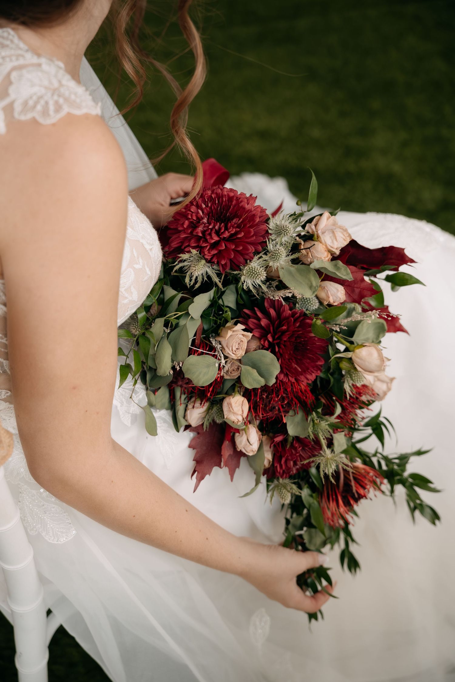 Bride in a white lace gown holds a burgundy and blush bridal bouquet of dahlias, spray roses and greenery on a lush green lawn.