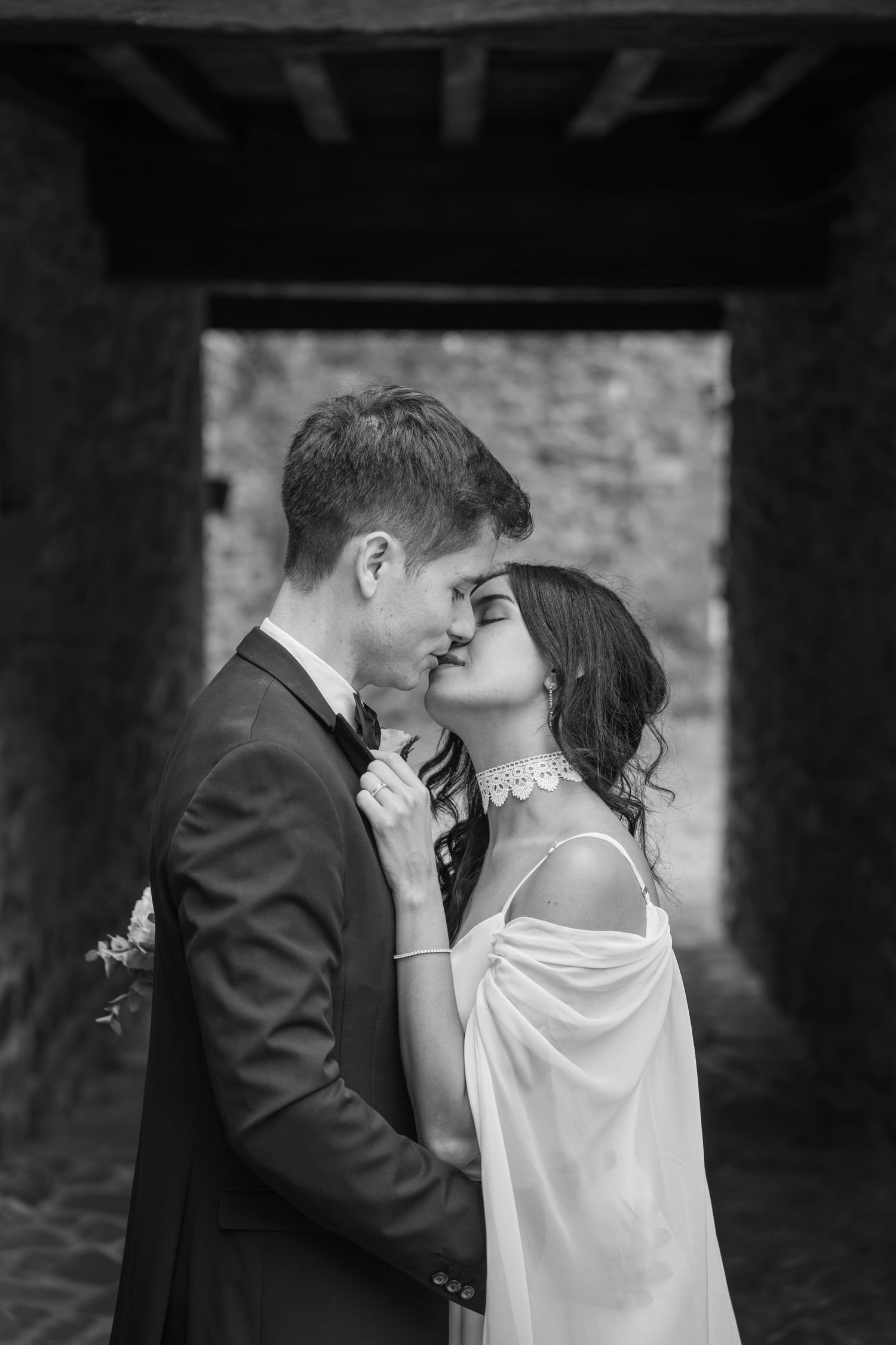 Black and white photo of a bride and groom sharing a kiss in a stone archway, she in a white shawl and pearl necklace, he in a tuxedo.