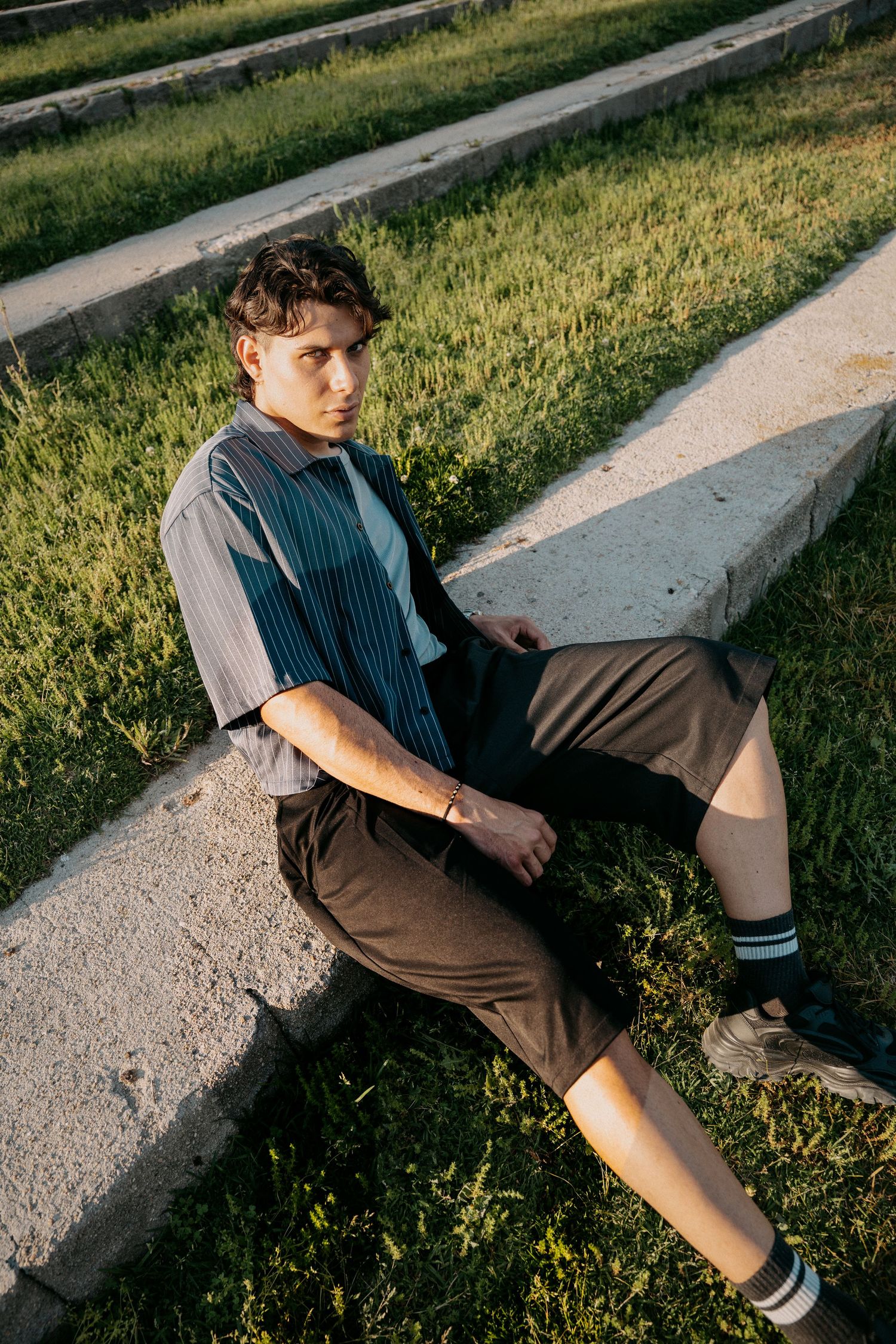 Young man in a striped button-up shirt, brown shorts and sneakers sits on a concrete step beside grass in warm evening sunlight.
